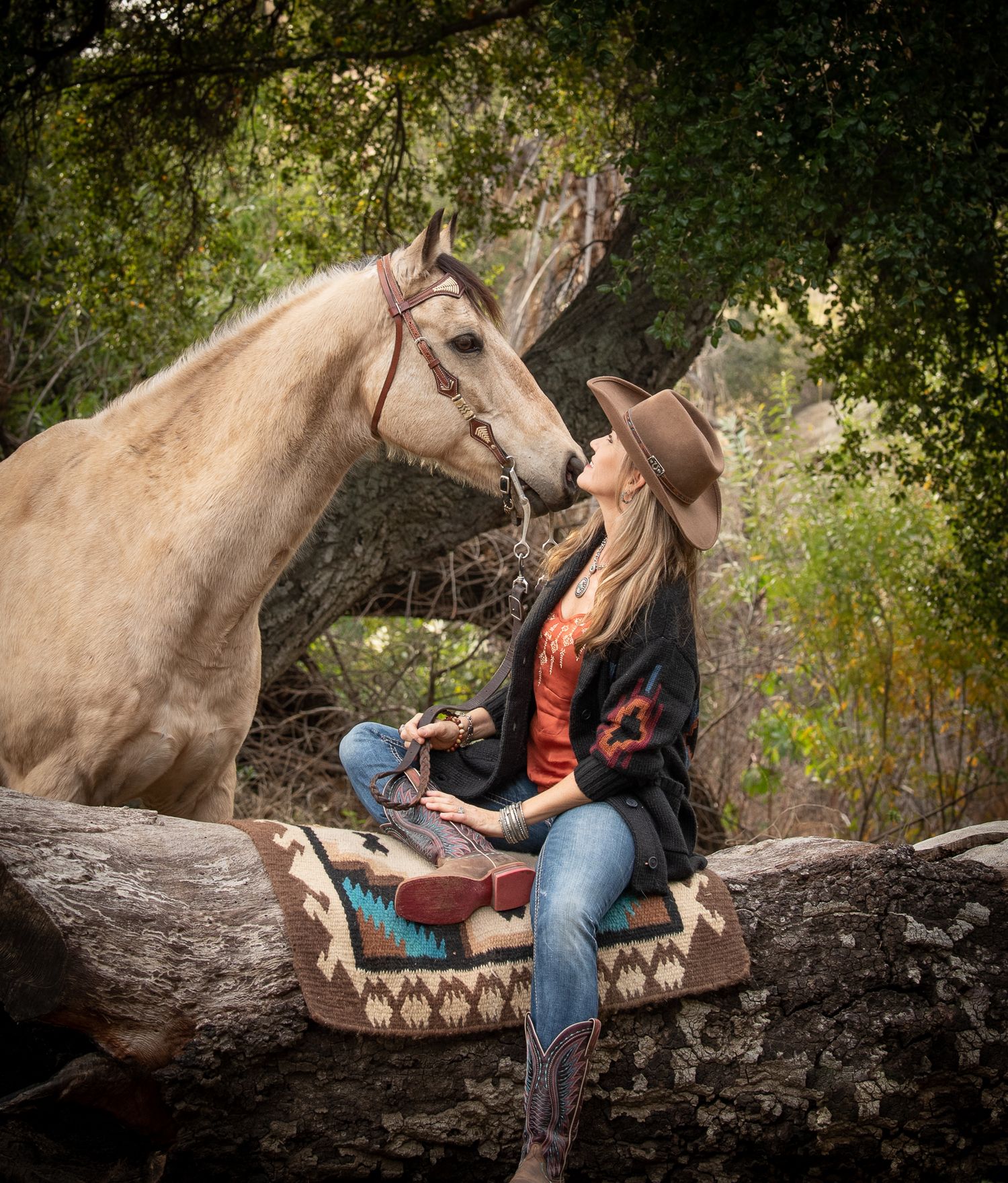 Blonde woman in cowboy hat sitting on log gazing up at palomino horse in forest.