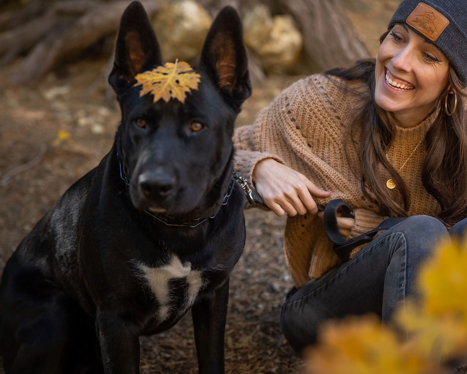 A black dog with a yellow autumn leaf on its head sits beside a smiling woman in a beanie outdoors.