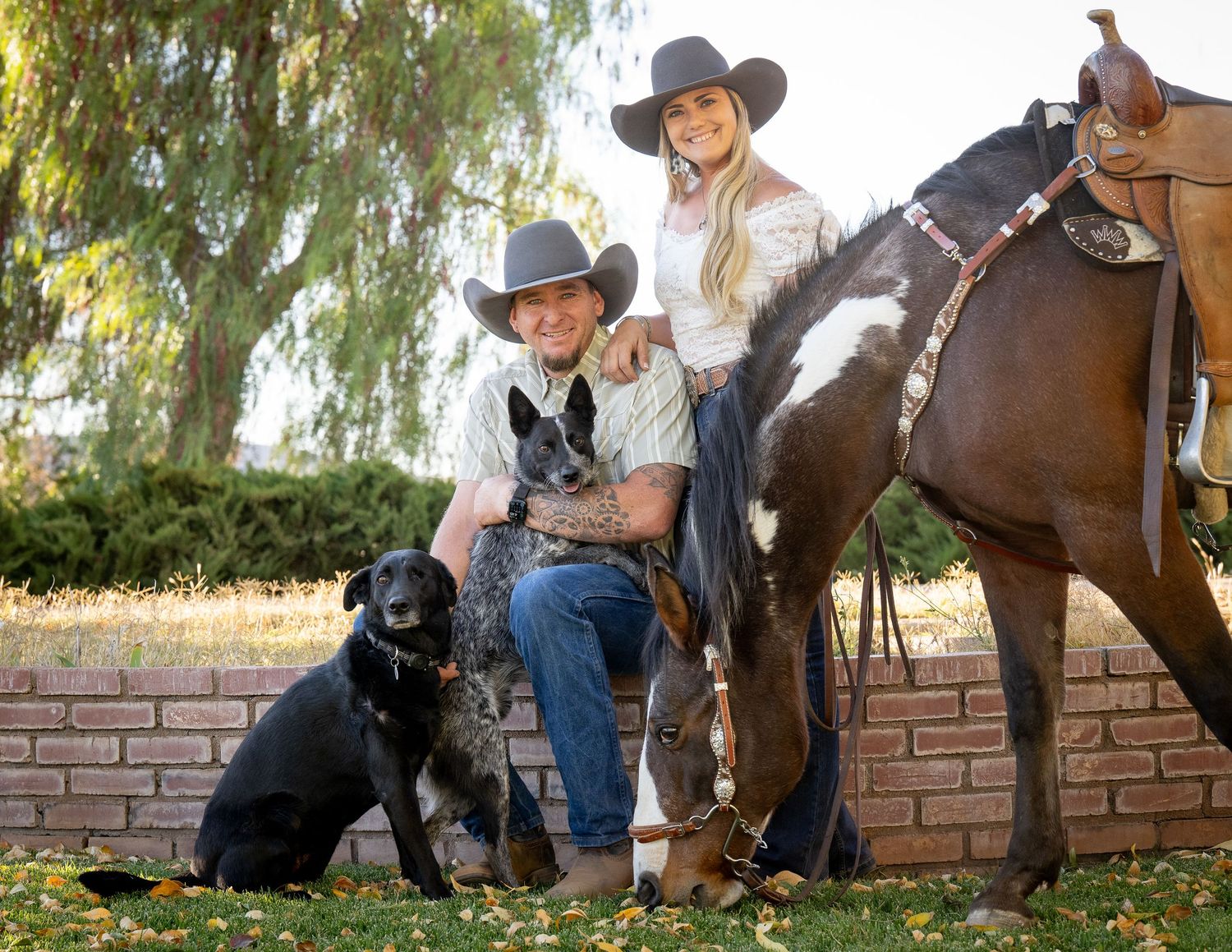 Couple in cowboy hats poses with two dogs and a paint horse beside a brick wall outdoors.