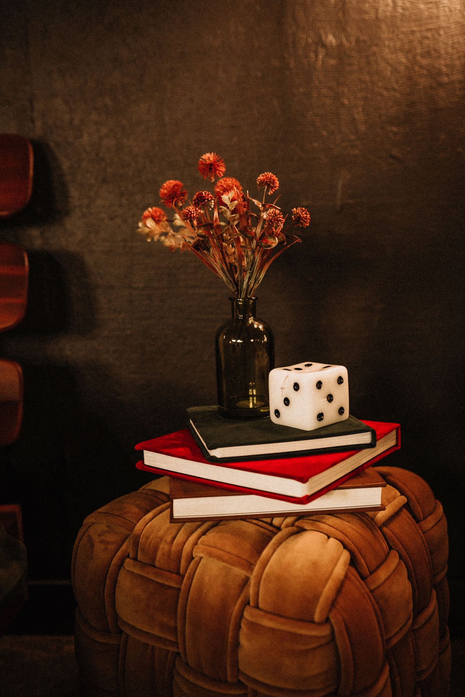 Still life with dried flowers, books, and decorative dice on a leather ottoman in moody lighting.