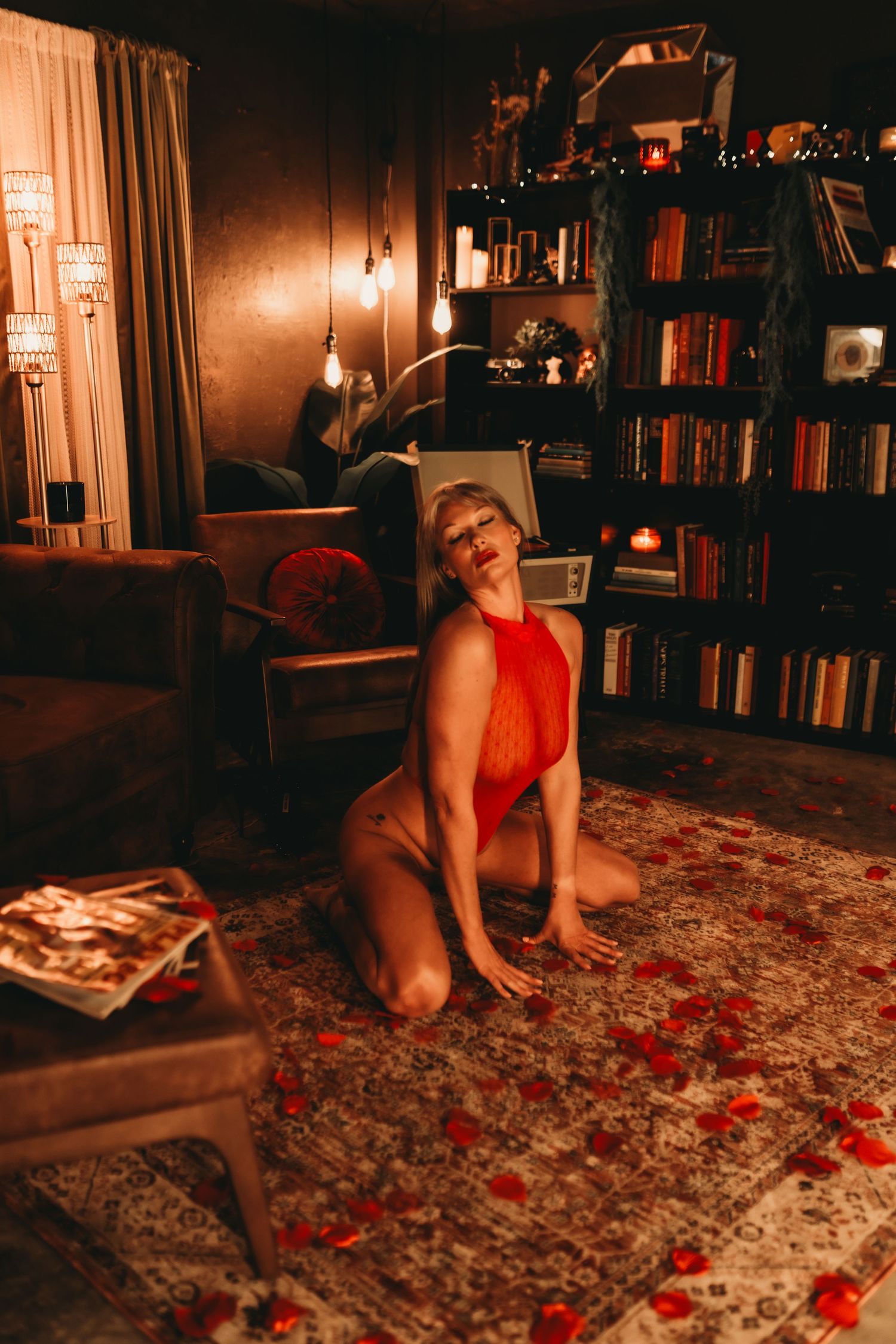 Woman in red bodysuit kneeling on a rug in a warmly lit room with bookshelves and rose petals.