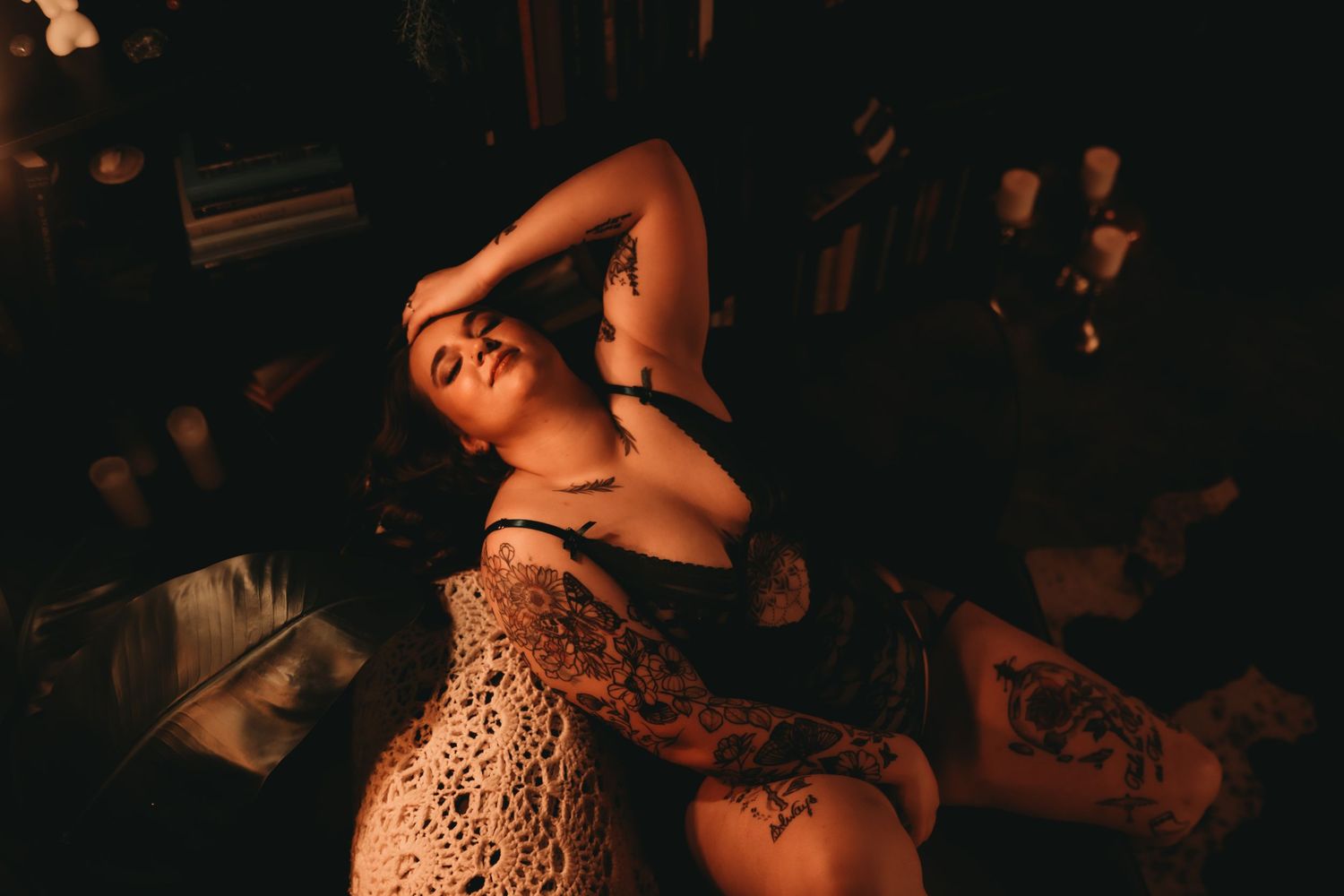 Woman with tattoos posing in dim warm lighting wearing lace and lingerie in a moody, atmospheric setting.