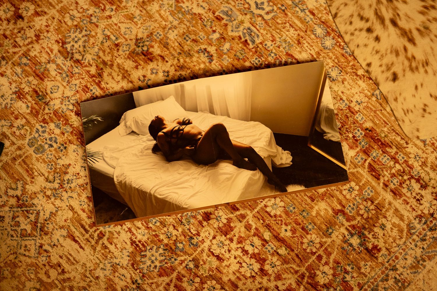 A person lying on a white bed, viewed through a mirror framed by an ornate golden patterned carpet.