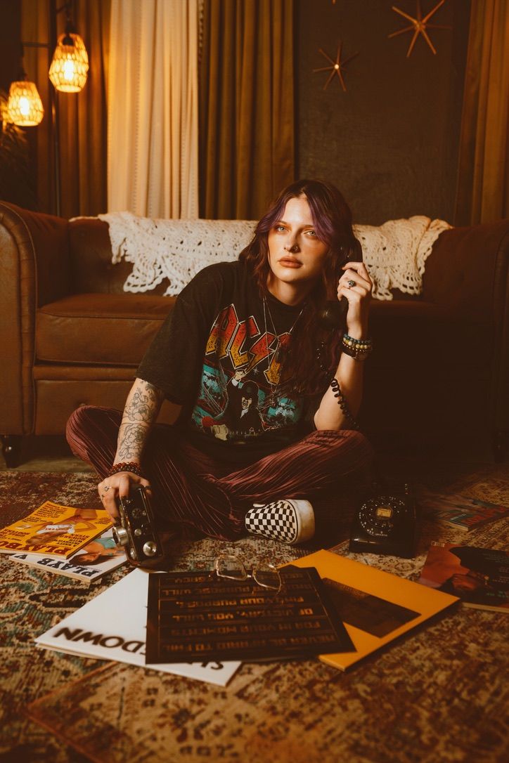 A person in gothic attire sits on a patterned carpet with a Ouija board in warm, moody lighting.