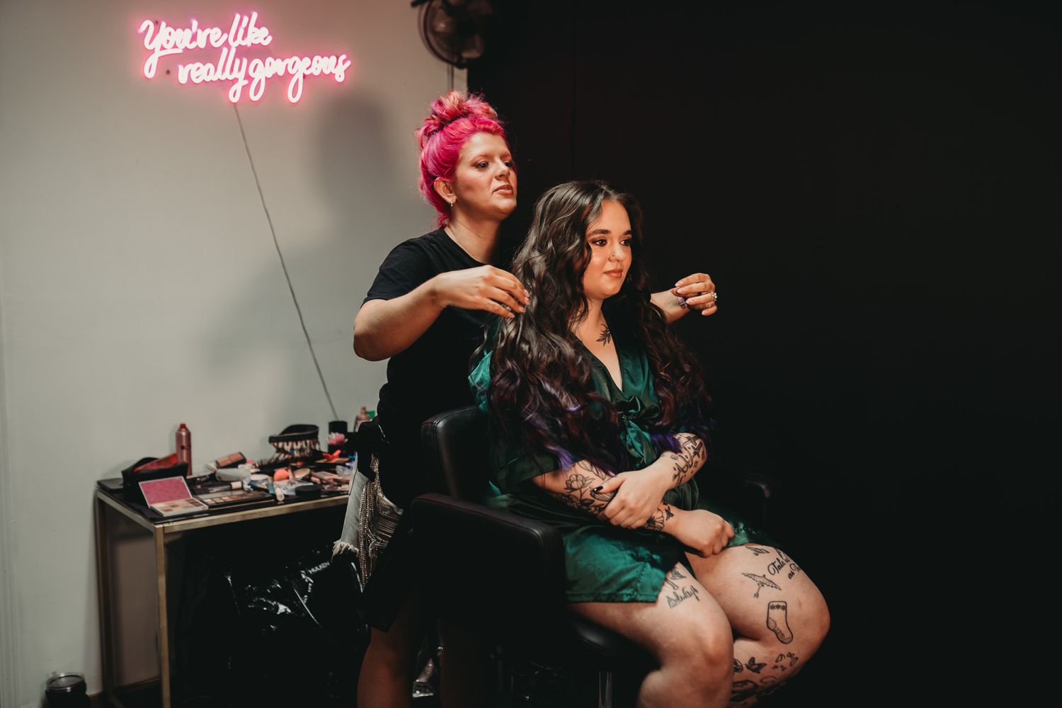 A pink-haired makeup artist applies makeup to a tattooed woman in a salon with neon sign decor.