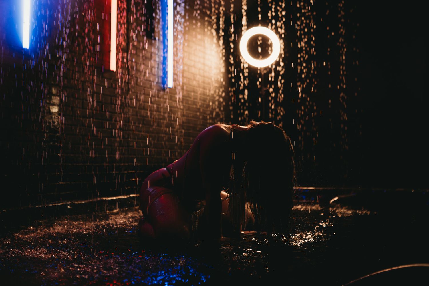 Person kneeling in rain at night with glowing ring light and neon signs in dark atmospheric setting.