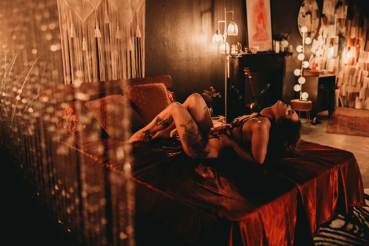 Woman reclining on a red velvet bed in a warmly lit bohemian room with decorative lights and macrame wall hangings.