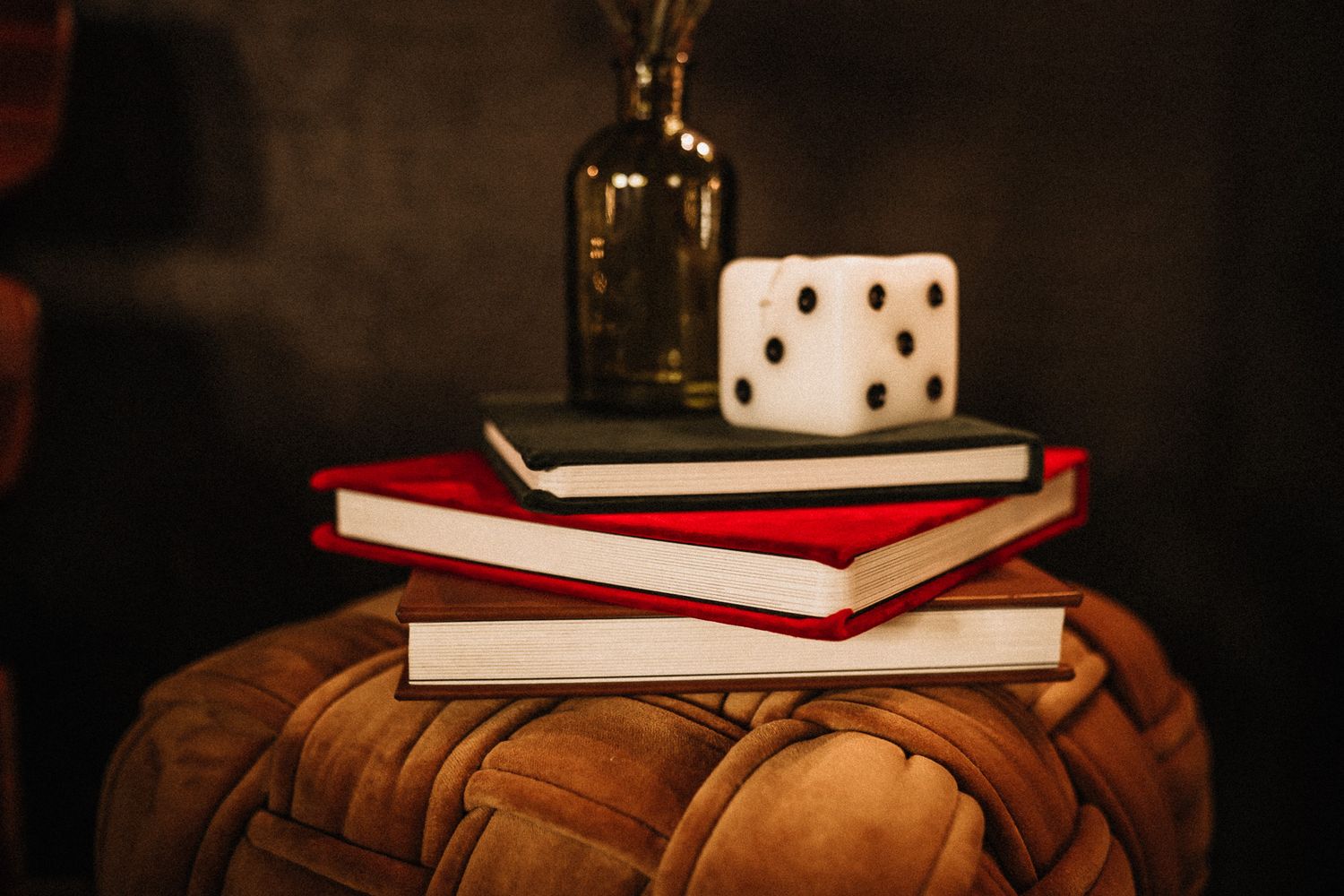 A large white decorative dice sits atop two stacked books on a brown leather pouf beside a glass vase.