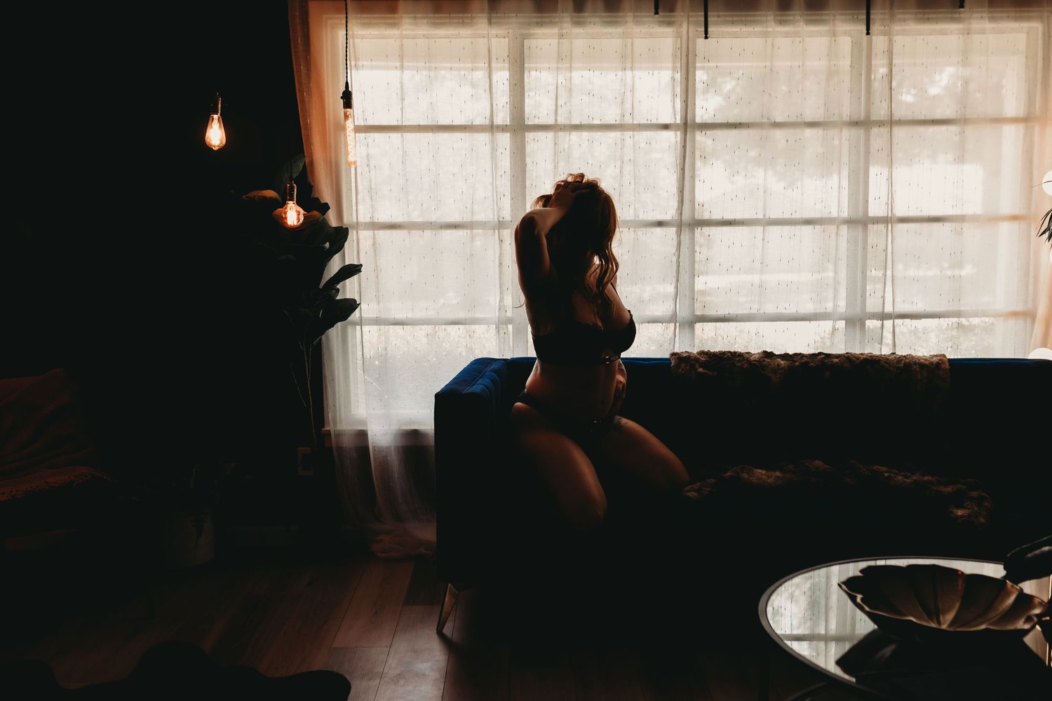 Silhouette of a woman sitting by a bright window in a moody, dimly lit living room with warm ambient lighting.