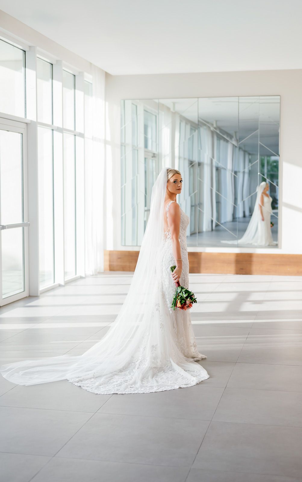 A bride in a flowing white wedding gown with a long cathedral veil poses in a bright modern hallway with floor-to-ceiling windows.