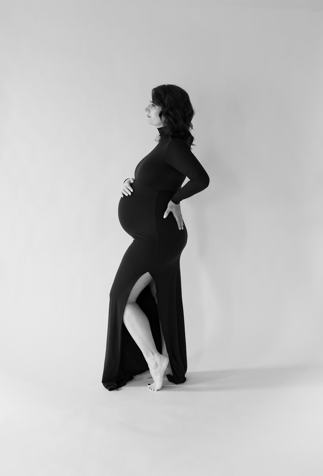 Artistic black and white maternity photo showing silhouette in long black dress with side slit against plain background.