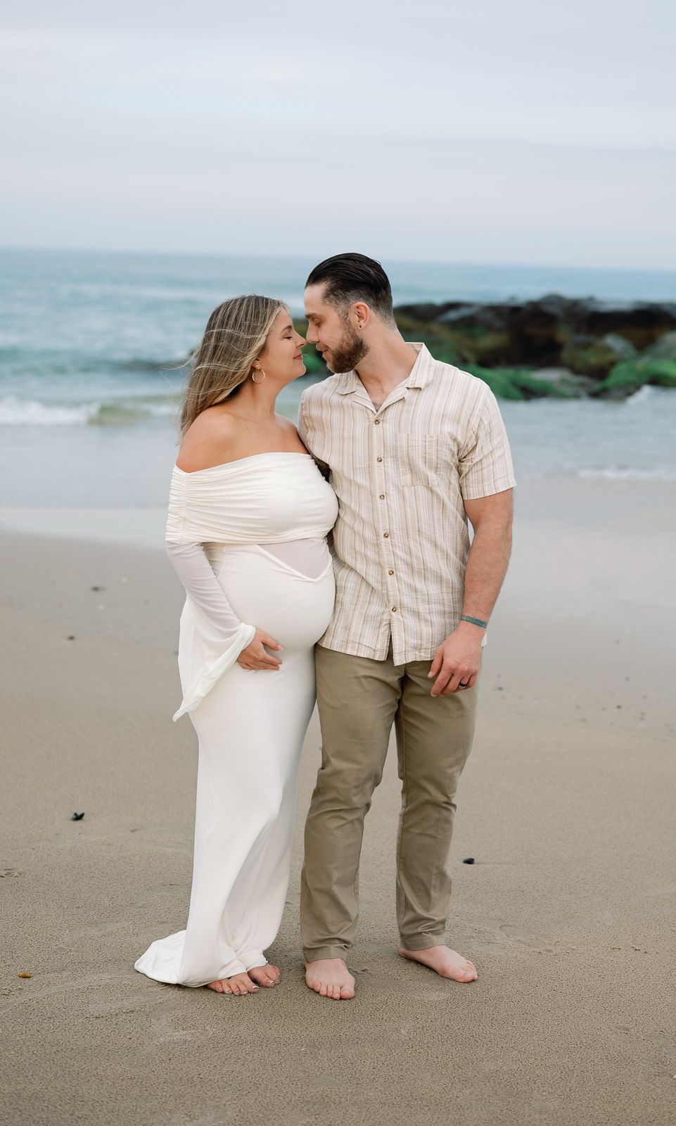 A NJ maternity couple shares an intimate moment on a misty beach wearing coordinated white and beige outfits.