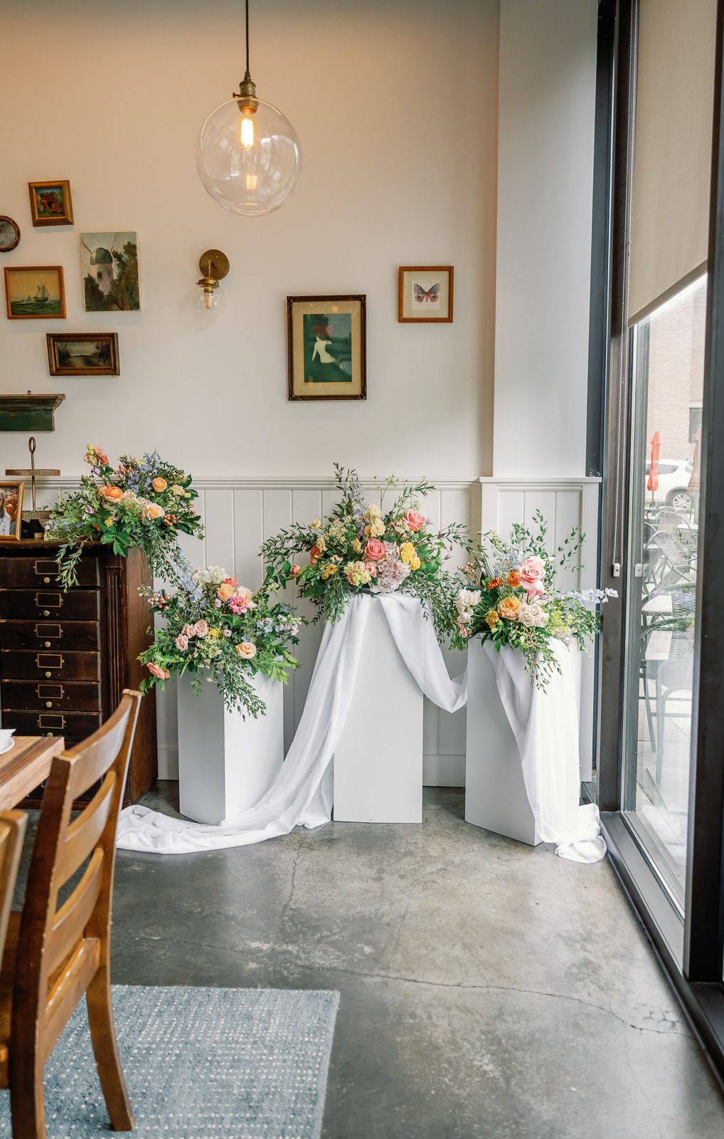 Modern interior space with concrete floors and floral arrangements on wooden pedestals near large windows.