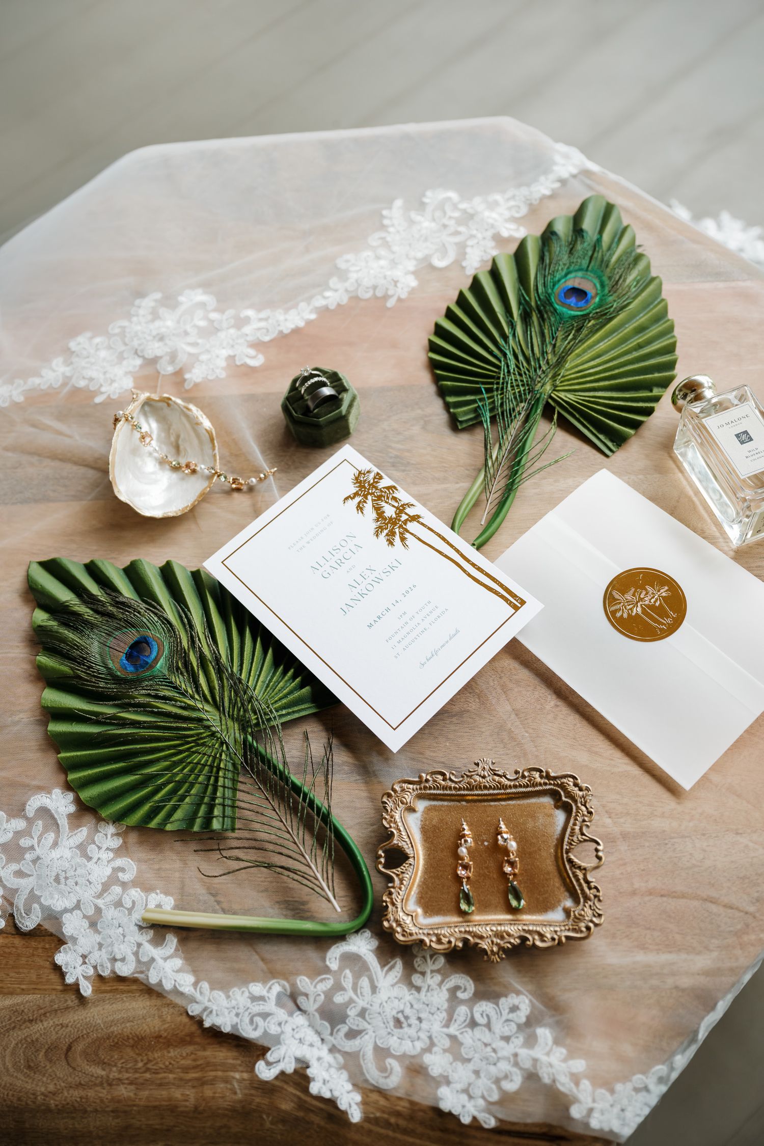 Wedding invitation flatlay with palm leaves, lace, gold frame, and delicate white stationery details on a wooden surface.