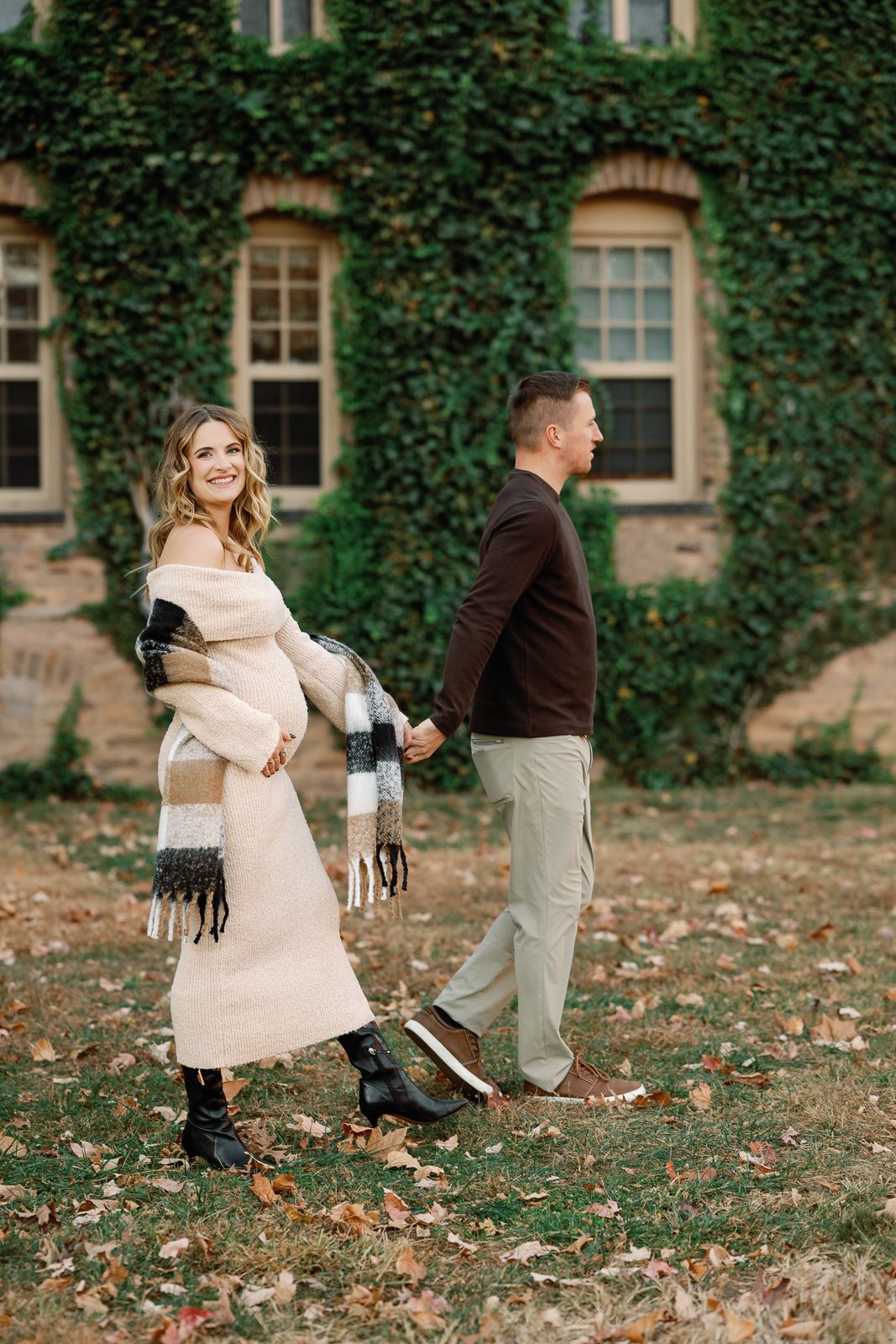 A couple in fall attire playfully poses for photos on fallen leaves near an ivy-covered stone building in NJ.