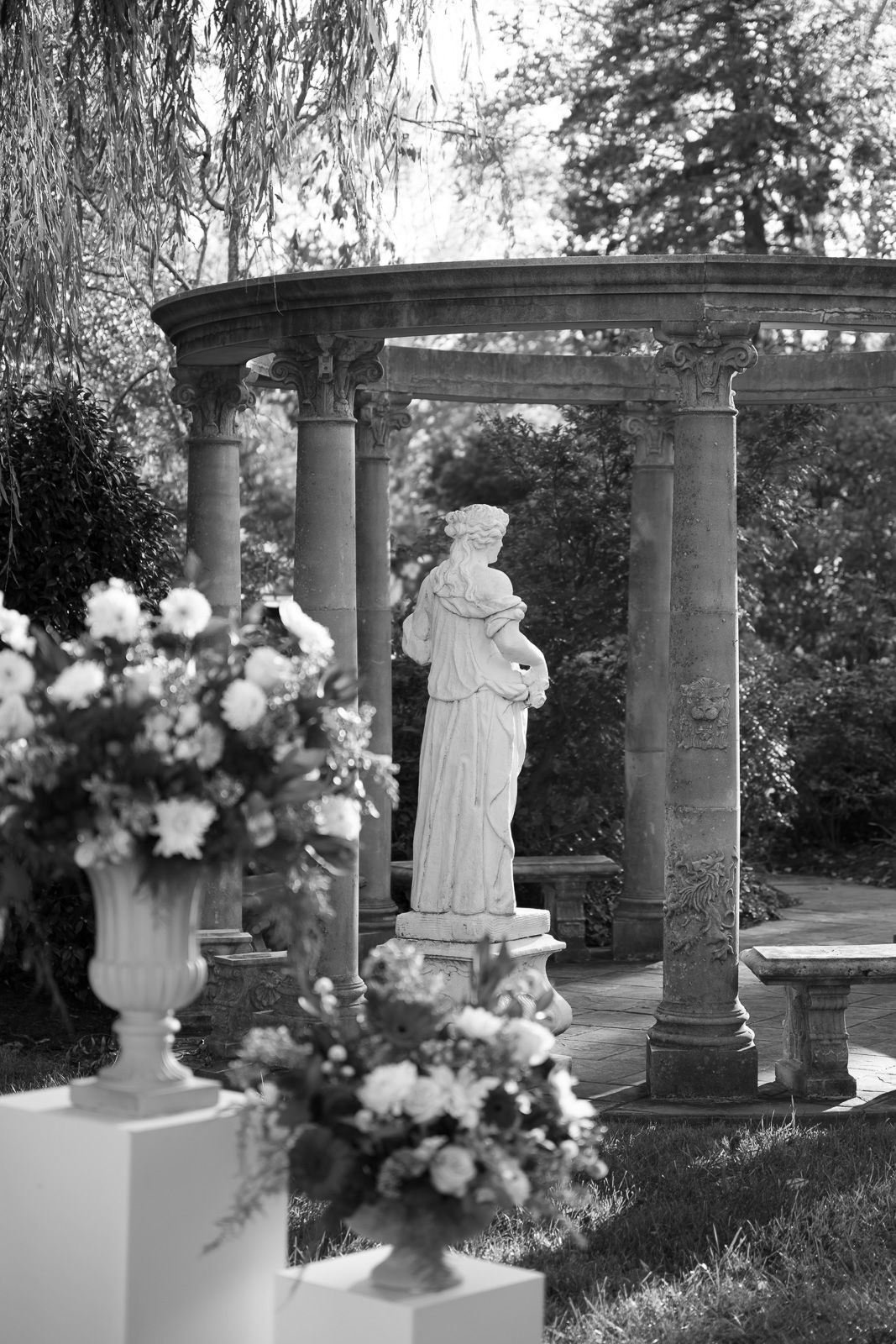 A classical garden setting with white stone columns and statues surrounded by floral arrangements and greenery.