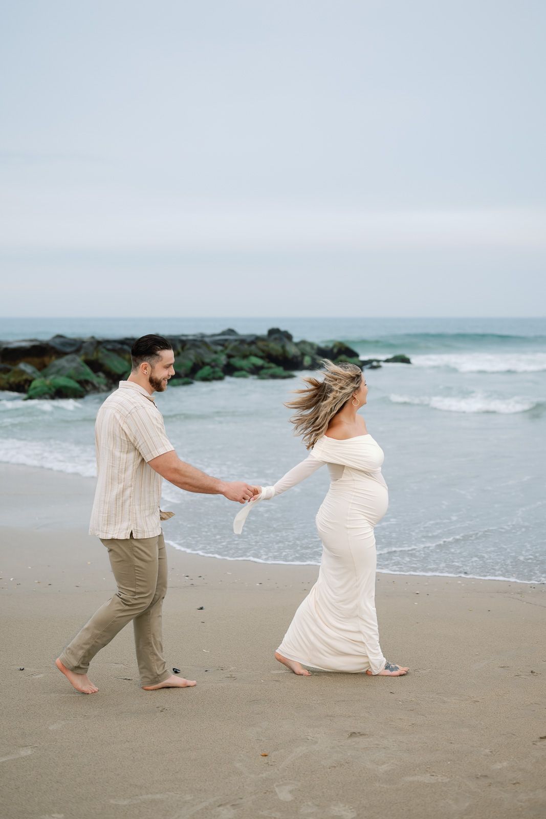 A New Jersey maternity photographer captures a couple in light colored clothing holding hands on a misty beach with waves in the background.