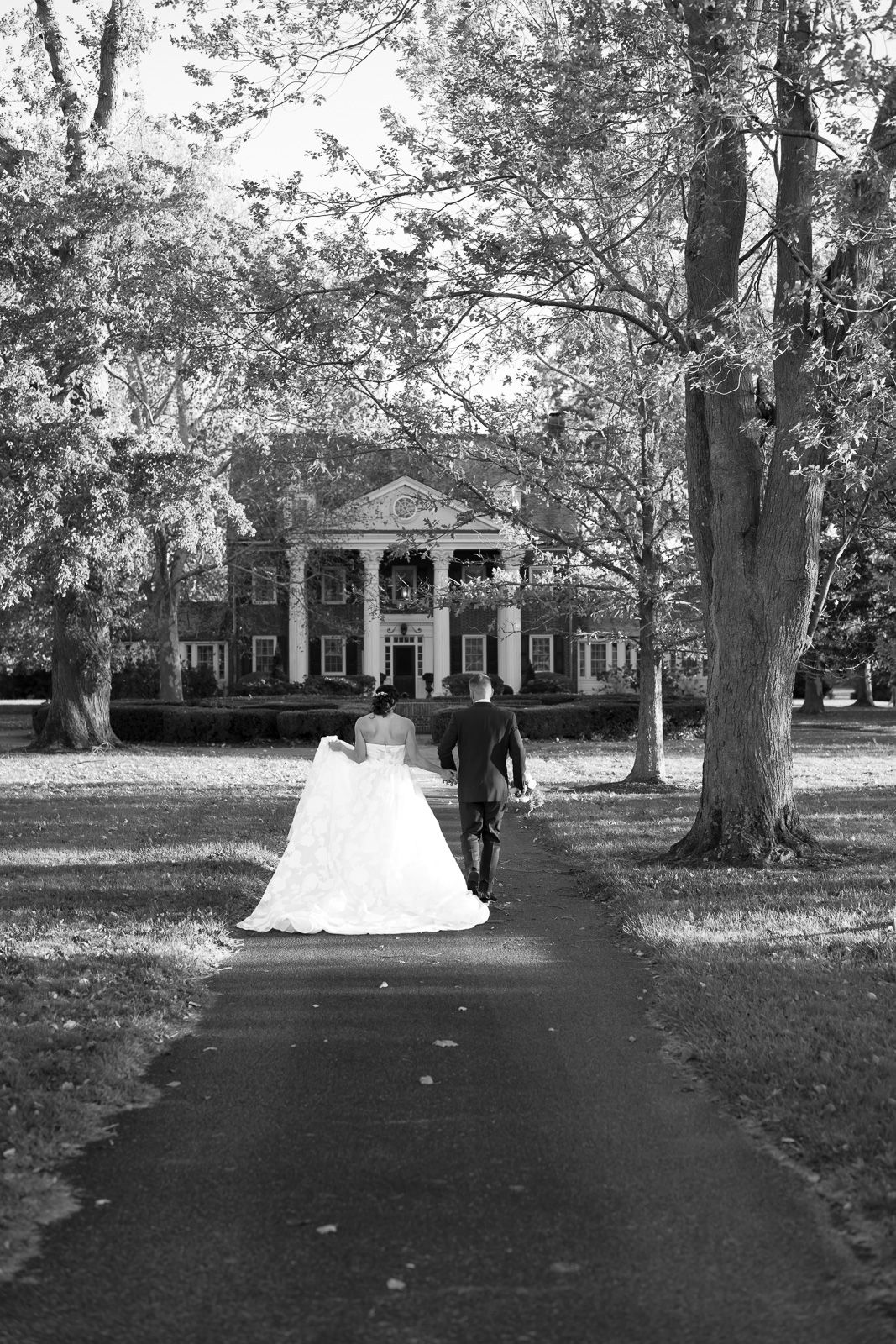 A couple walks down a tree-lined path towards a grand Southern mansion with white columns in black and white photography.