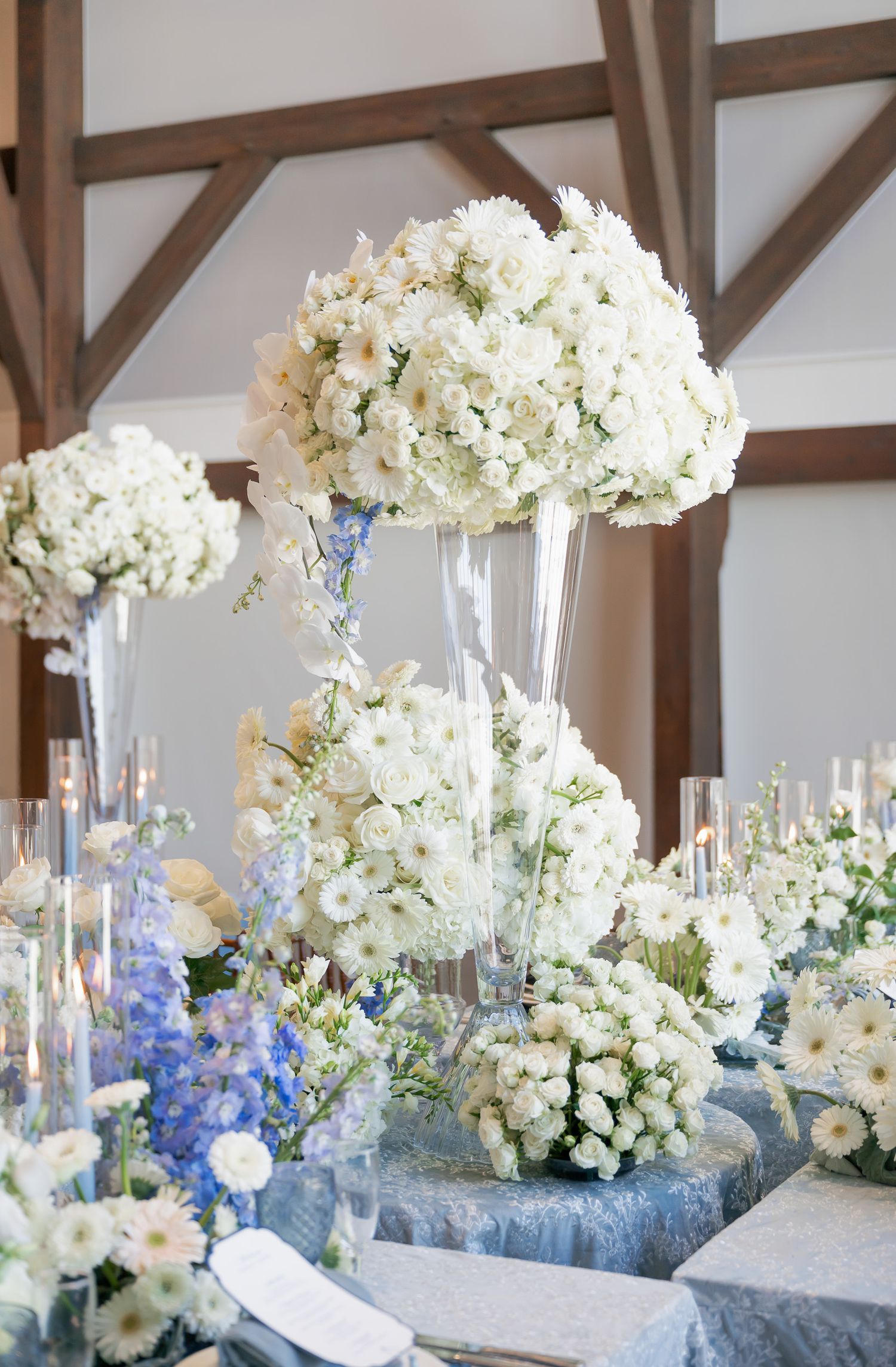 Elegant wedding table centerpieces feature tall glass vases with white hydrangeas and blue delphinium flowers.