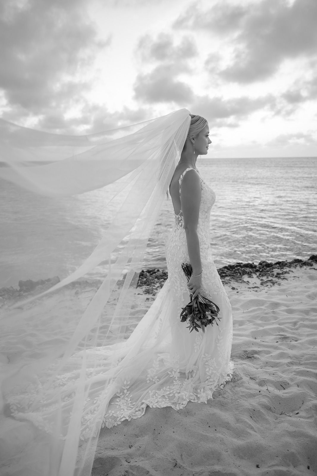 A flowing white wedding dress billows in the ocean breeze as waves crash on a rocky beach at sunset.