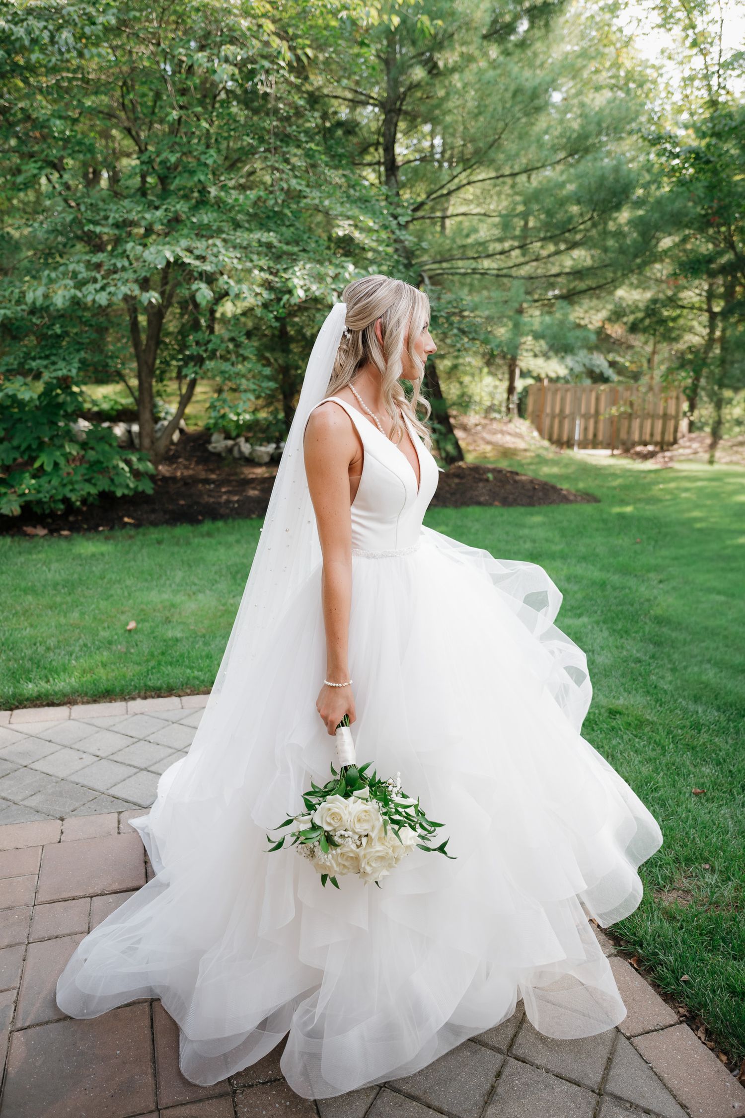 A flowing white wedding dress with layers of tulle fabric billows in the breeze on a stone patio surrounded by greenery.