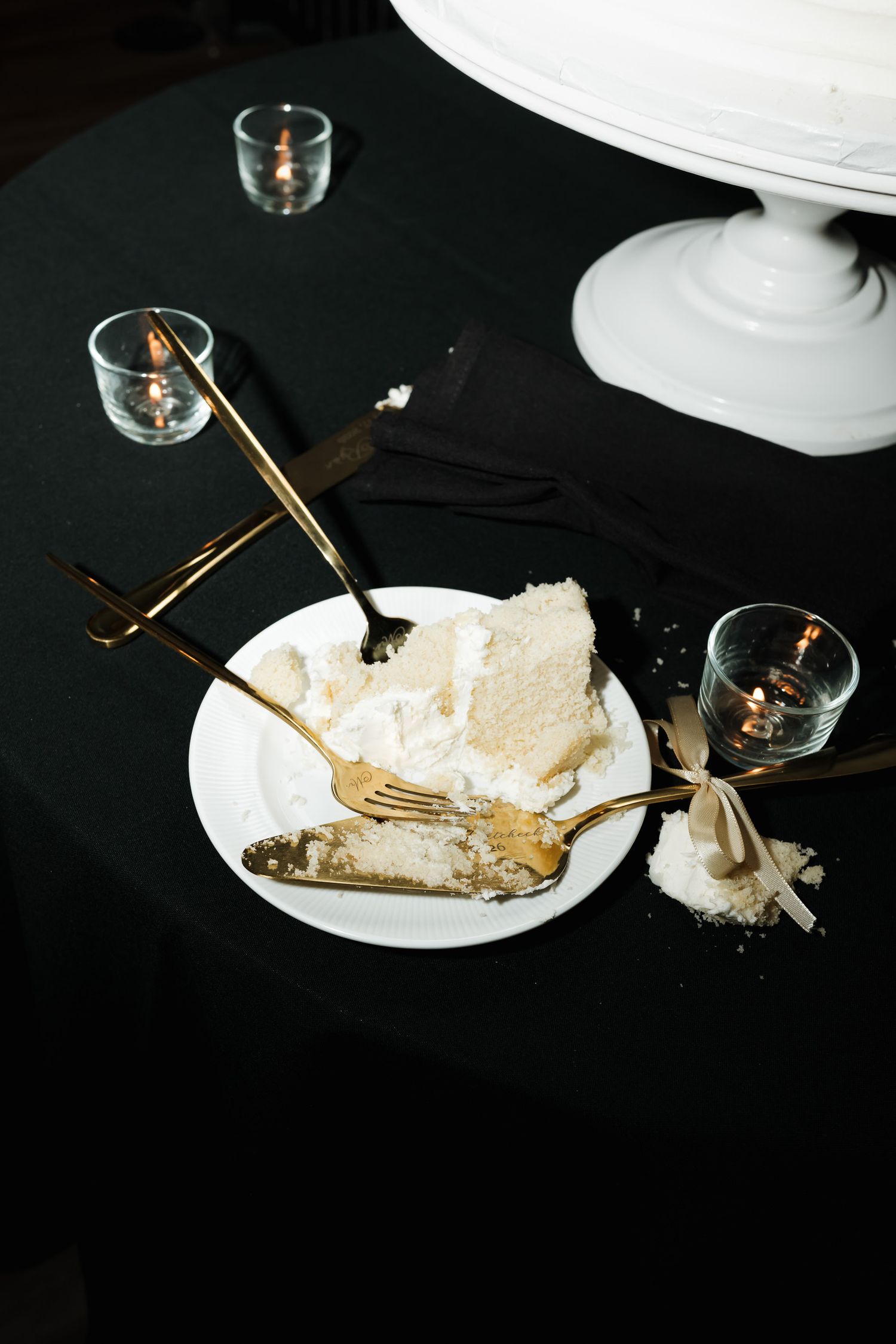 A slice of white cream pie with gold utensils on a dark table setting with small glasses.