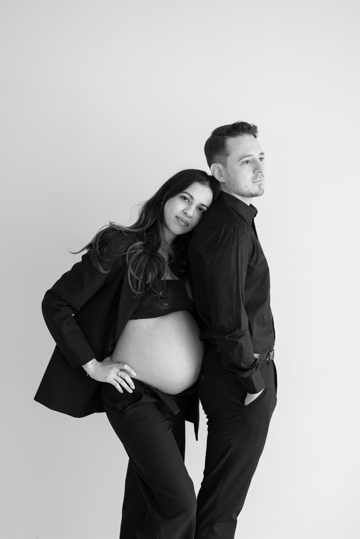 Black and white maternity photo of a couple in dark clothing posing together against a light background.