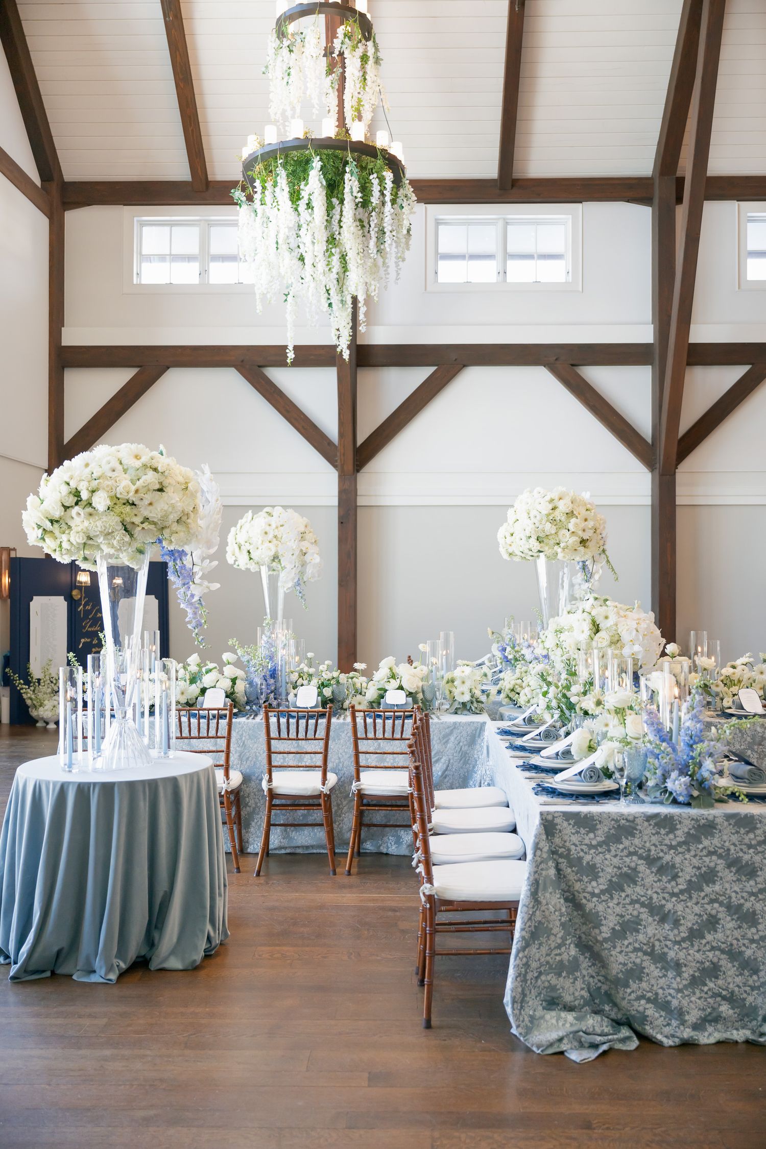 Elegant wedding reception venue with light blue tablecloths and white floral centerpieces in a rustic barn setting.