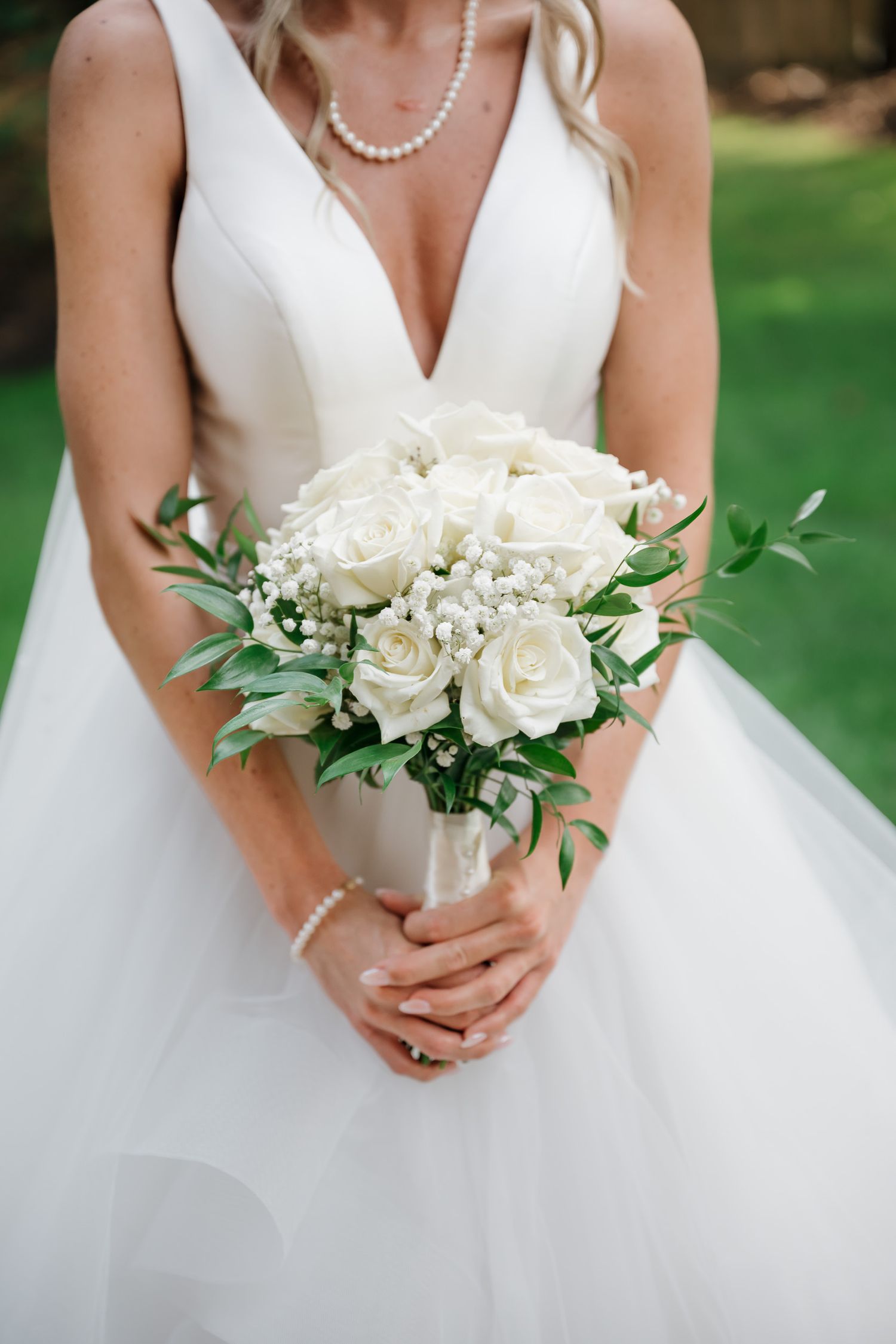 Close-up view of white rose bridal bouquet with greenery and pearl necklace details against outdoor garden background.