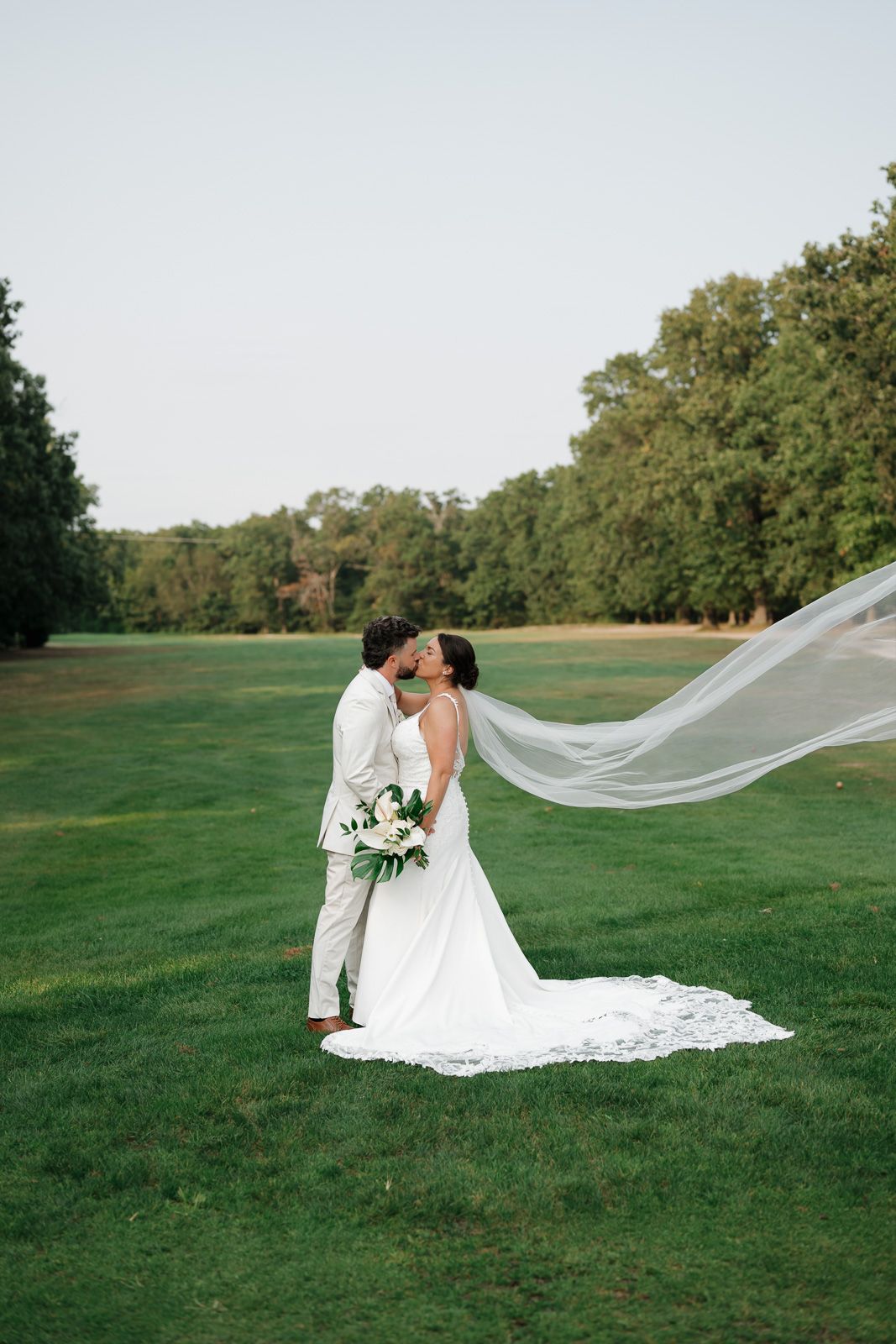 A wedding couple embraces on a lush green lawn with a flowing veil caught in the breeze at a NJ wedding venue.