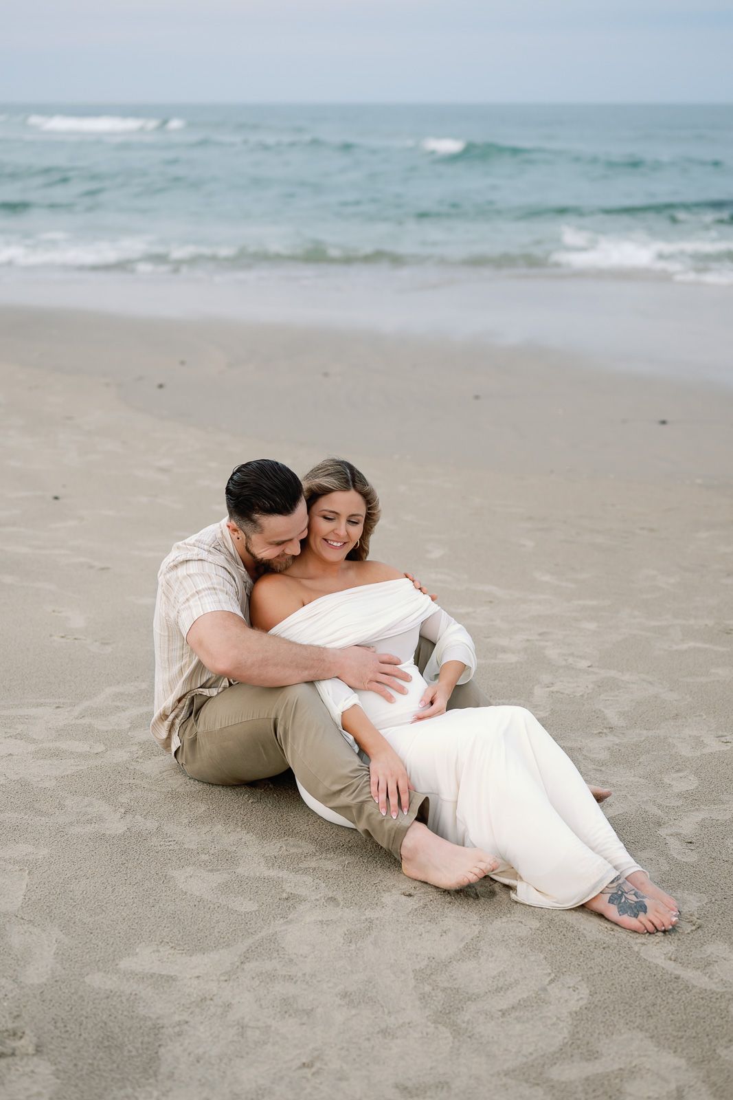 A couple embraces while sitting on a sandy beach with ocean waves in the background photographed by a NJ maternity photographer.