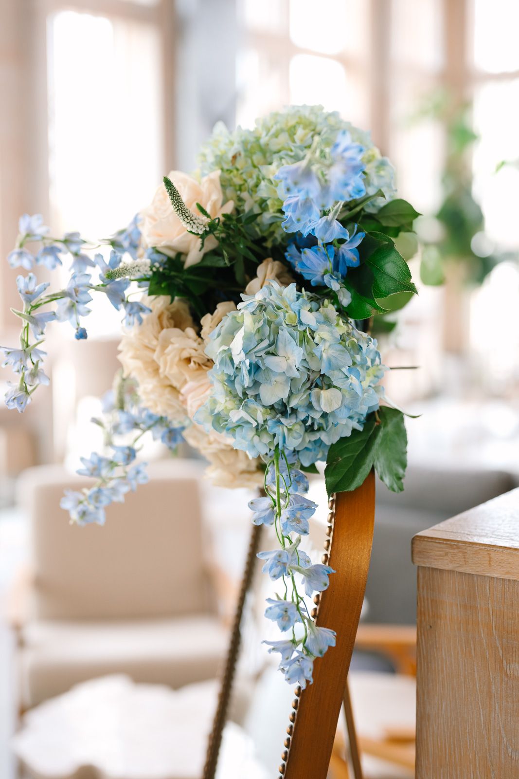 Light blue hydrangeas and cream colored flowers decorate wooden chairs in an elegant indoor New Jersey venue.