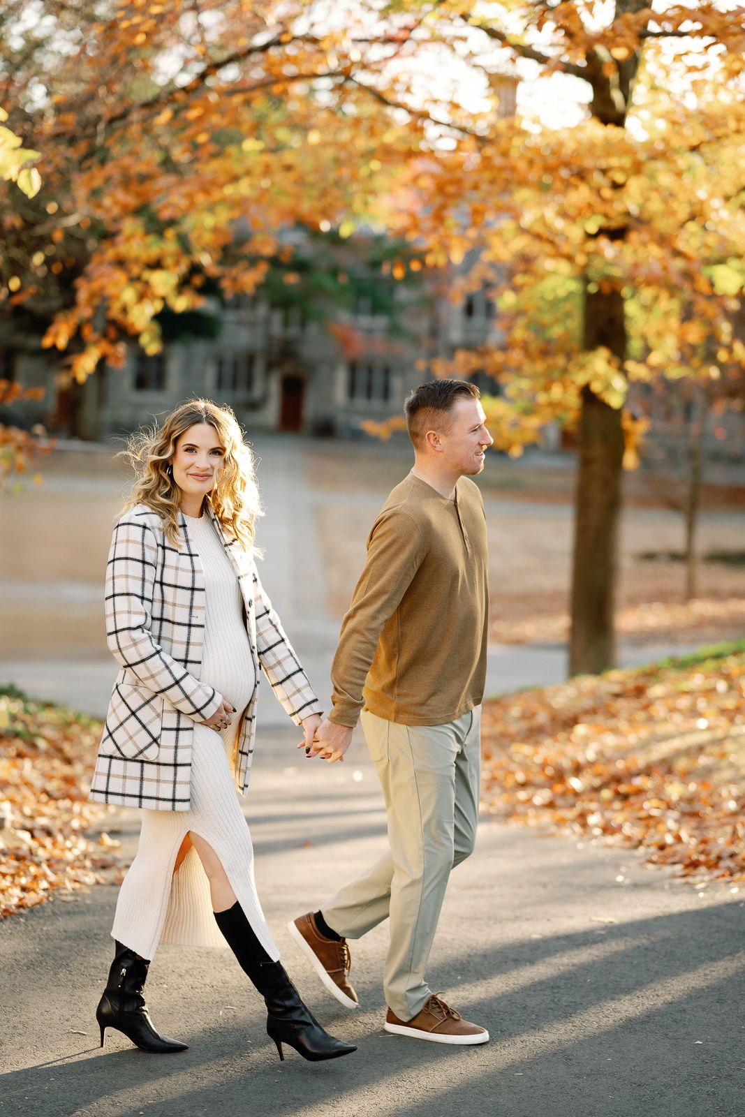 A NJ maternity photographer captures a couple playfully walking through fallen autumn leaves on a path lined with golden trees.