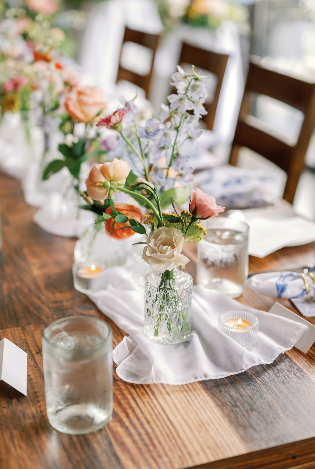 Small glass vases with delicate pink flowers and greenery line a rustic wooden table with white linens and tea lights.