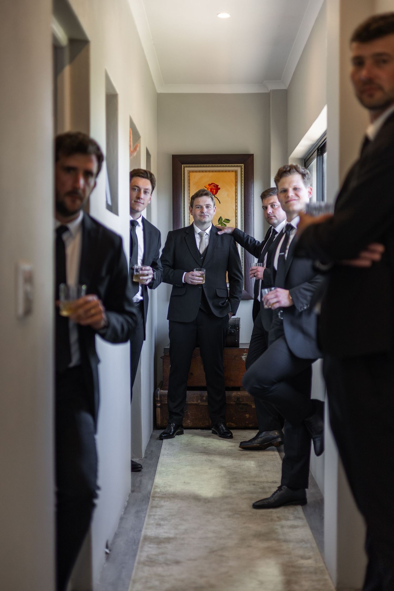 Group of groomsmen in black suits posing in hallway holding champagne glasses.