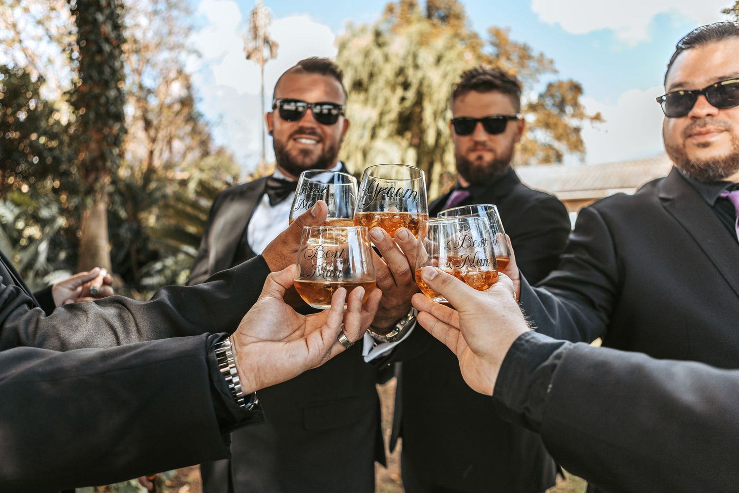 Groomsmen toasting with drinks while wearing sunglasses outdoors.