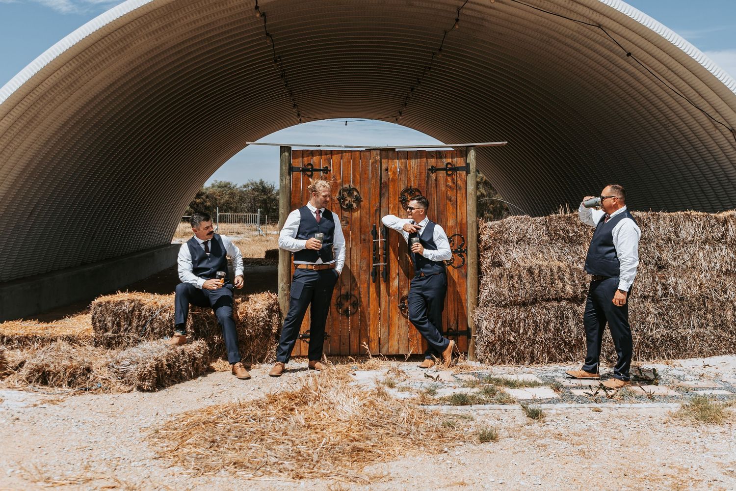 Wedding party poses together near rustic hay bales and metal barn structure on sunny day.