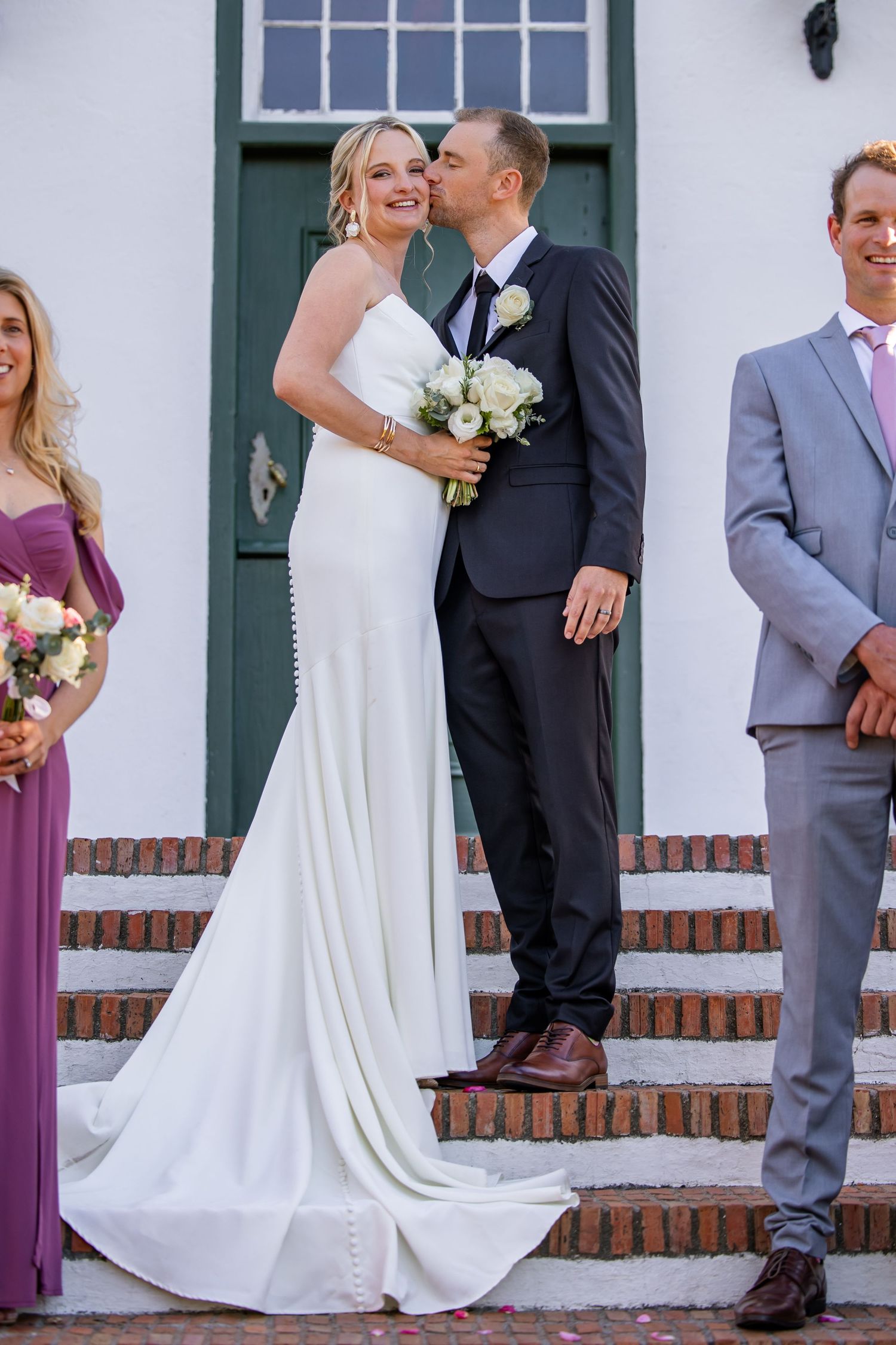 Wedding party poses on white steps with bridesmaids in purple dresses and groomsmen in gray suits.