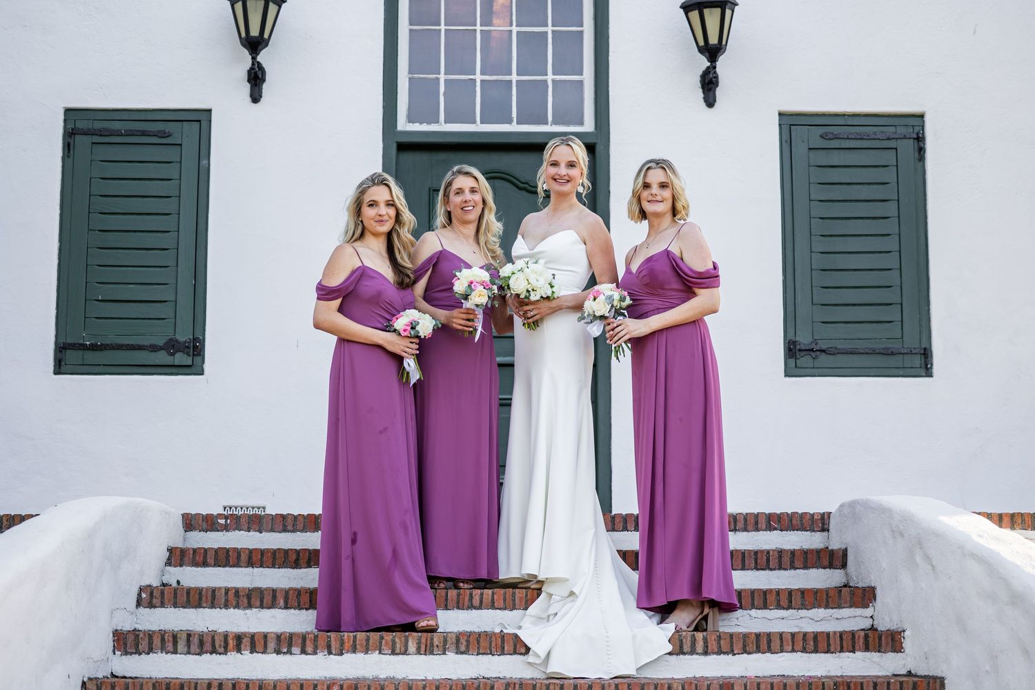 Bridal party in purple dresses posing on white steps with colonial architecture.