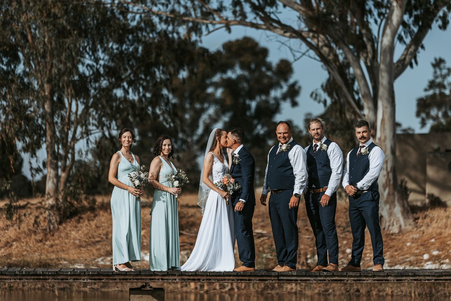 Wedding party in light blue bridesmaid dresses and dark suits poses together on wooden boardwalk near trees.