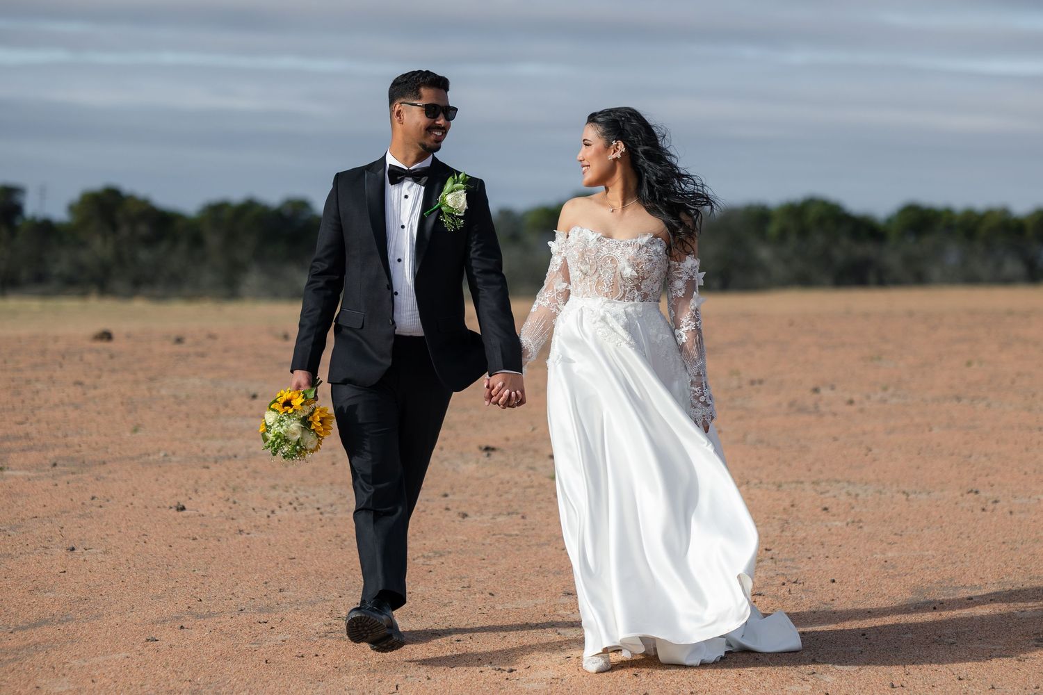 Newlywed couple walking hand-in-hand across red desert landscape, groom in tuxedo, bride in white lace off-shoulder gown.