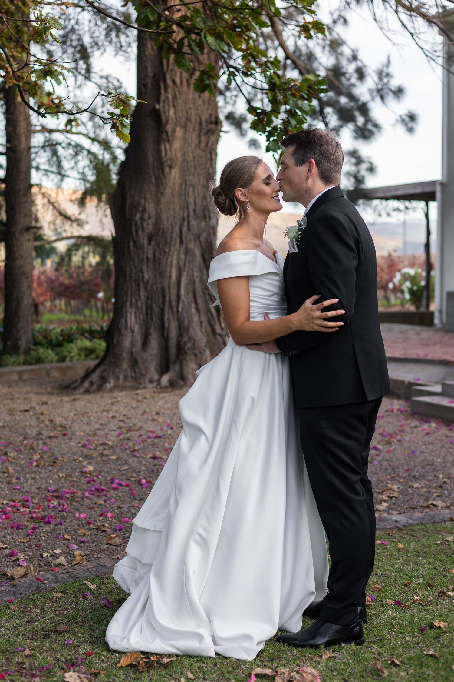 Bride and groom sharing a romantic kiss outdoors under a large tree, bride in off-shoulder white gown.