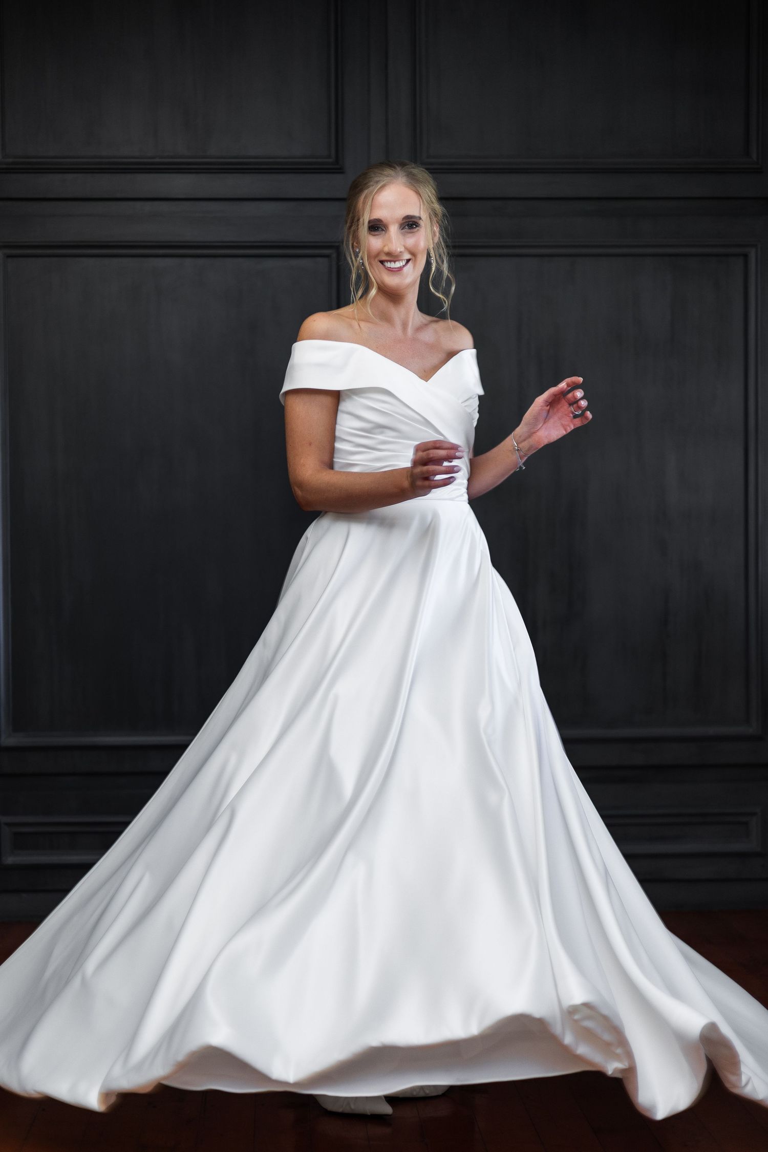 Radiant bride twirling in an elegant off-shoulder white satin ball gown against a dark paneled wall.