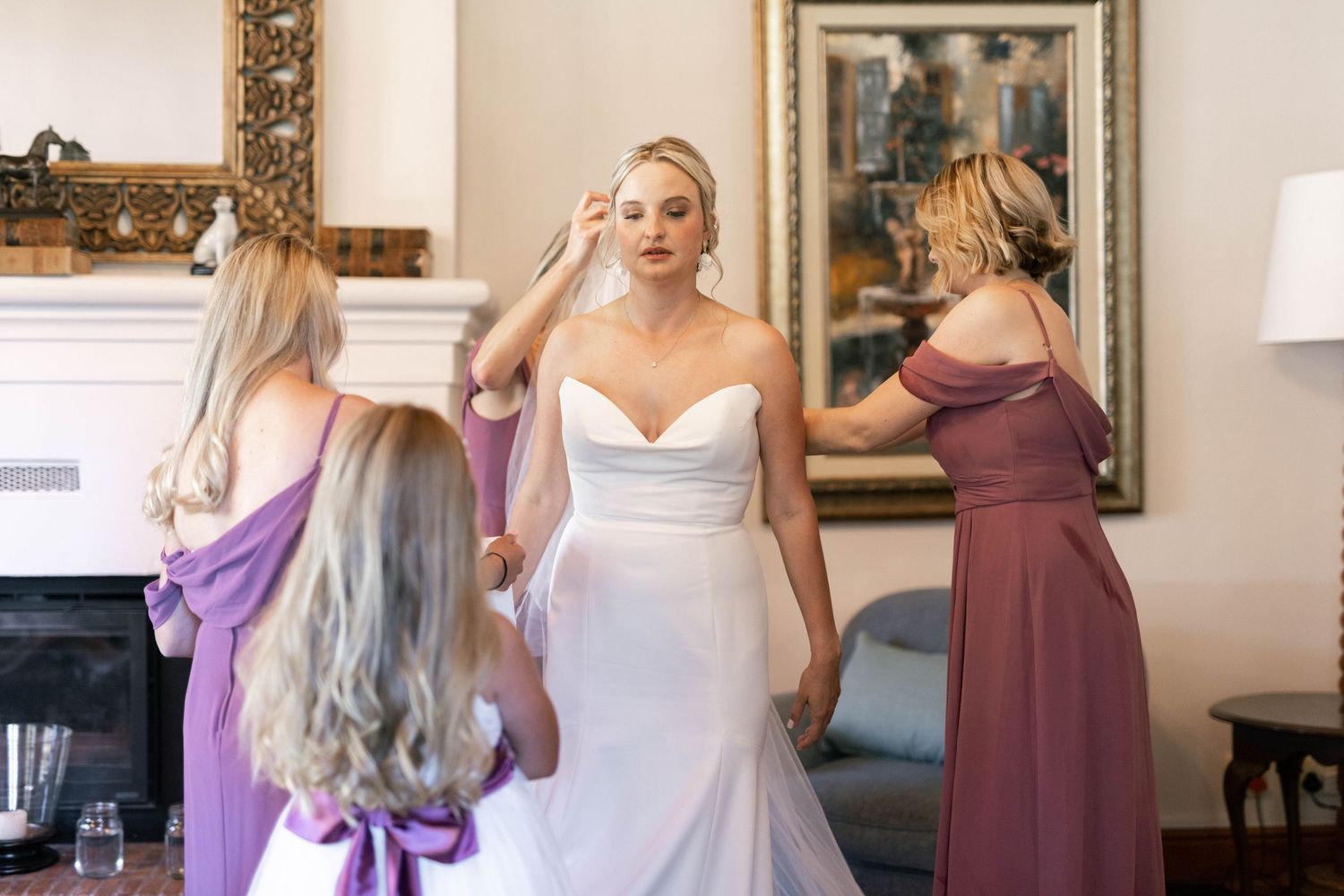 Wedding party getting ready in a room with bridesmaids in mauve dresses and bride in white gown.
