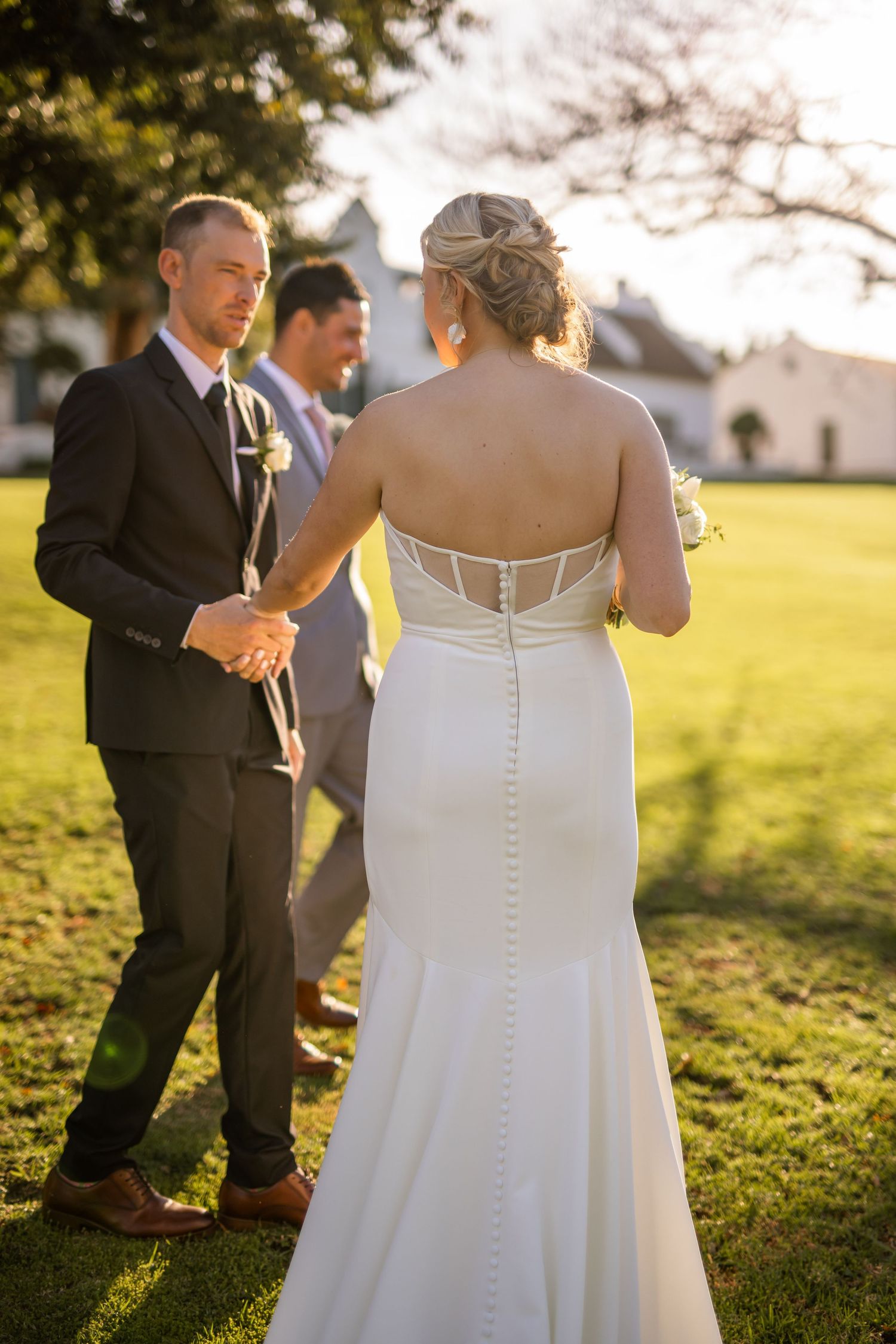 Wedding party gathered on sunny lawn at golden hour with elegant white gown details.