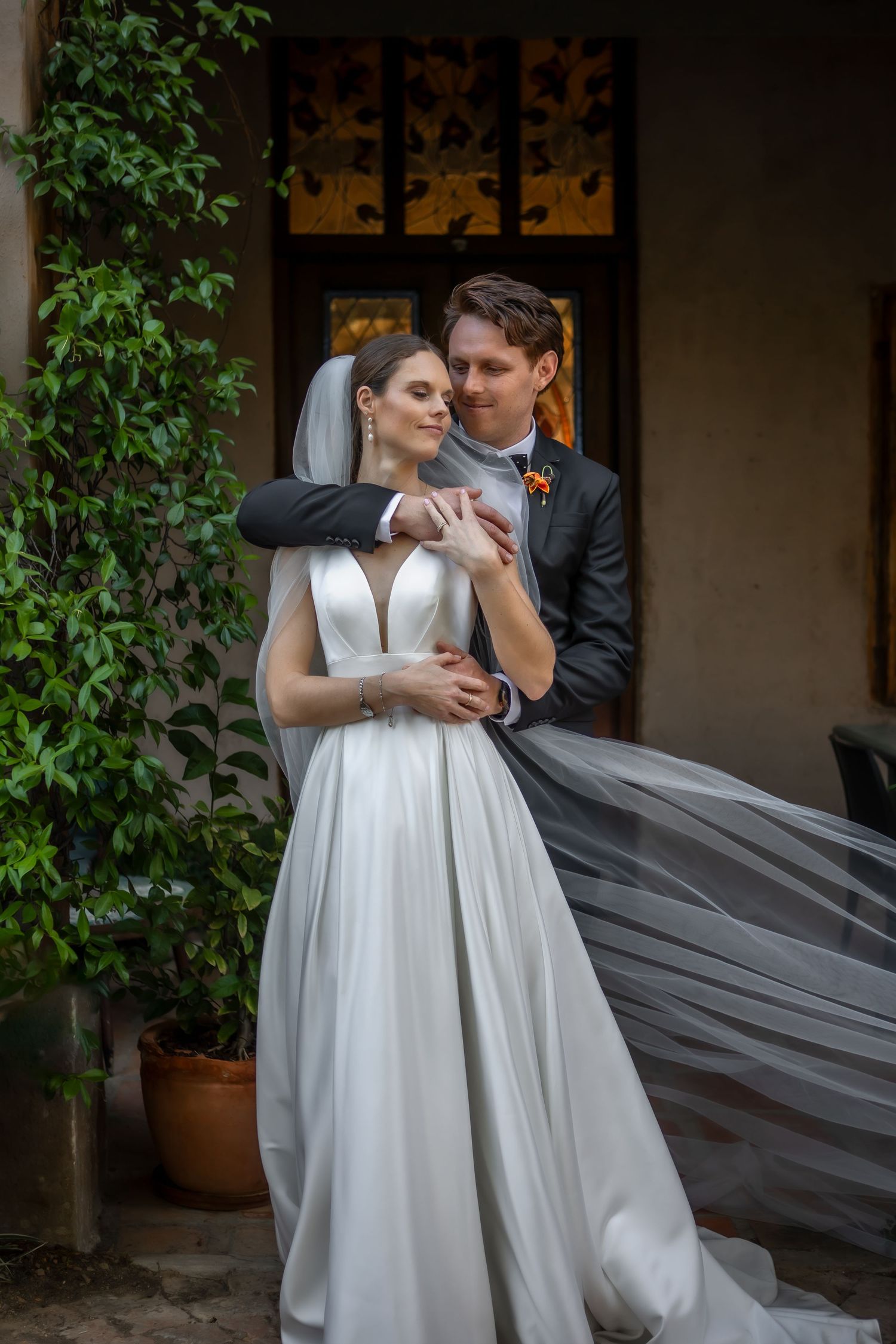 An elegant couple poses against a wall with decorative panels and climbing vines during their wedding.