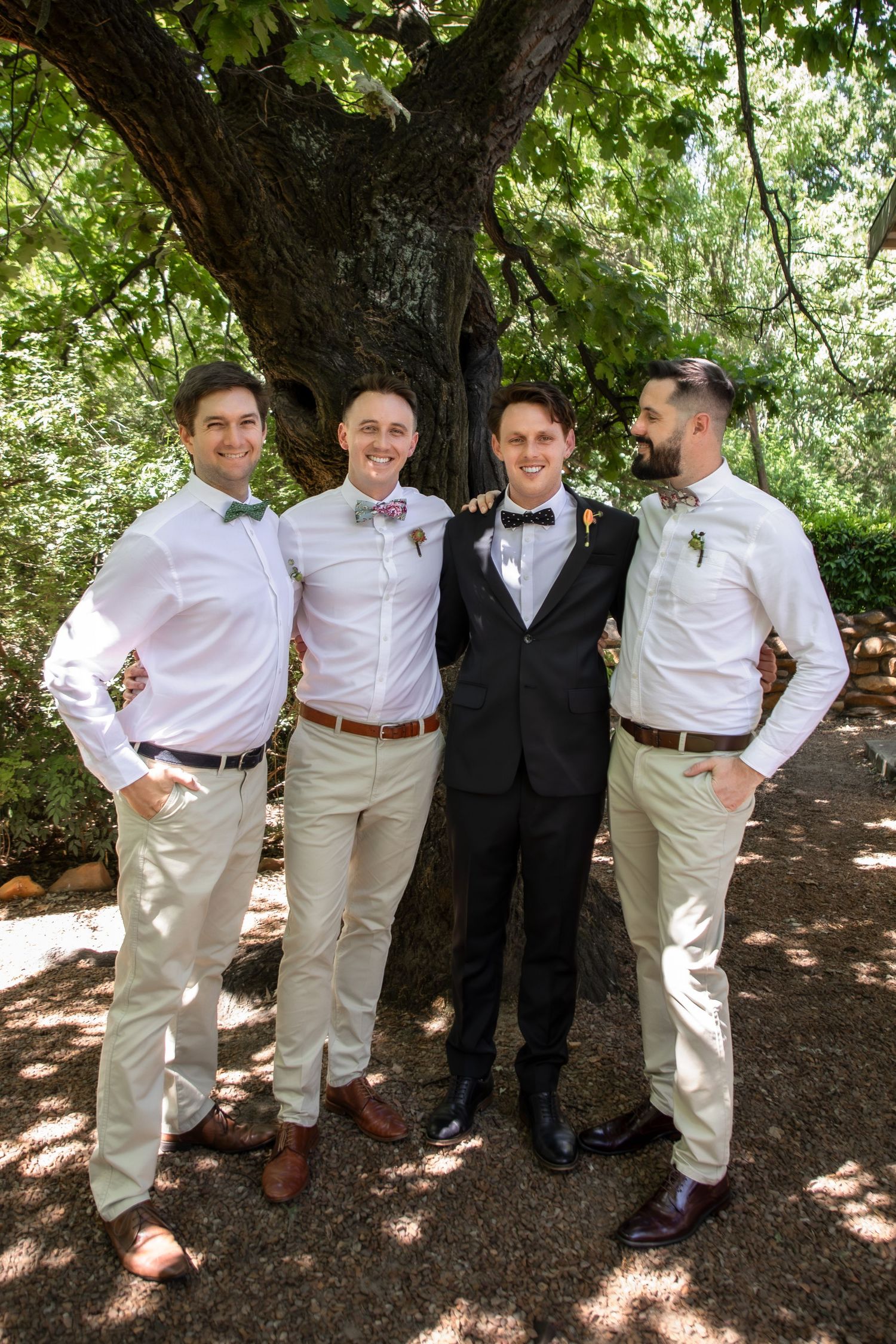 Groomsmen in white shirts and khaki pants pose casually under a shady tree.