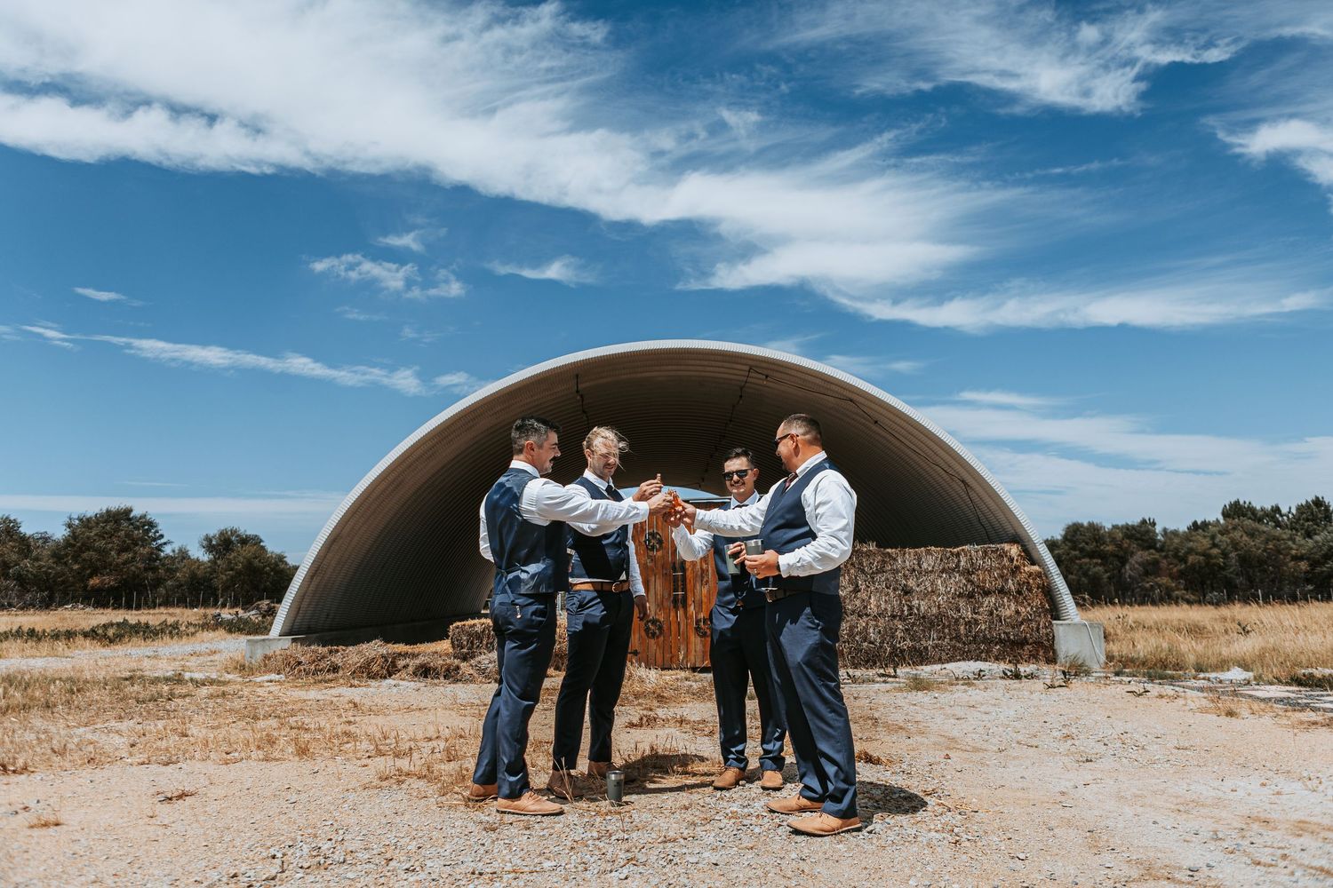 Wedding celebration group poses under large metal quonset hut against bright blue sky.