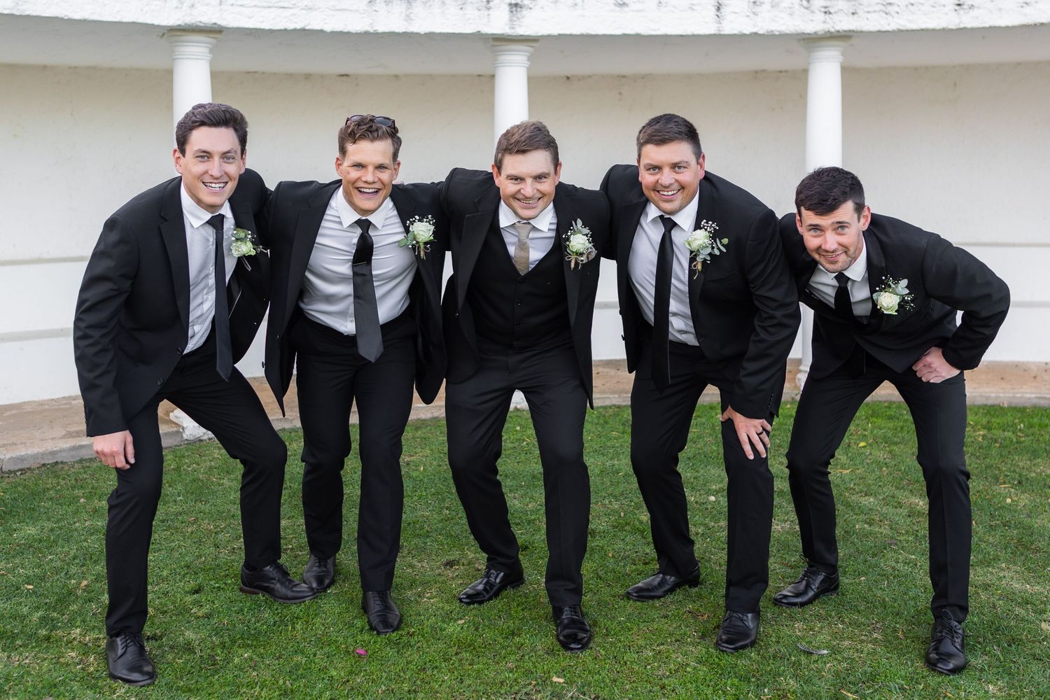 Groom and four groomsmen in black suits pose playfully together outdoors at a wedding venue.