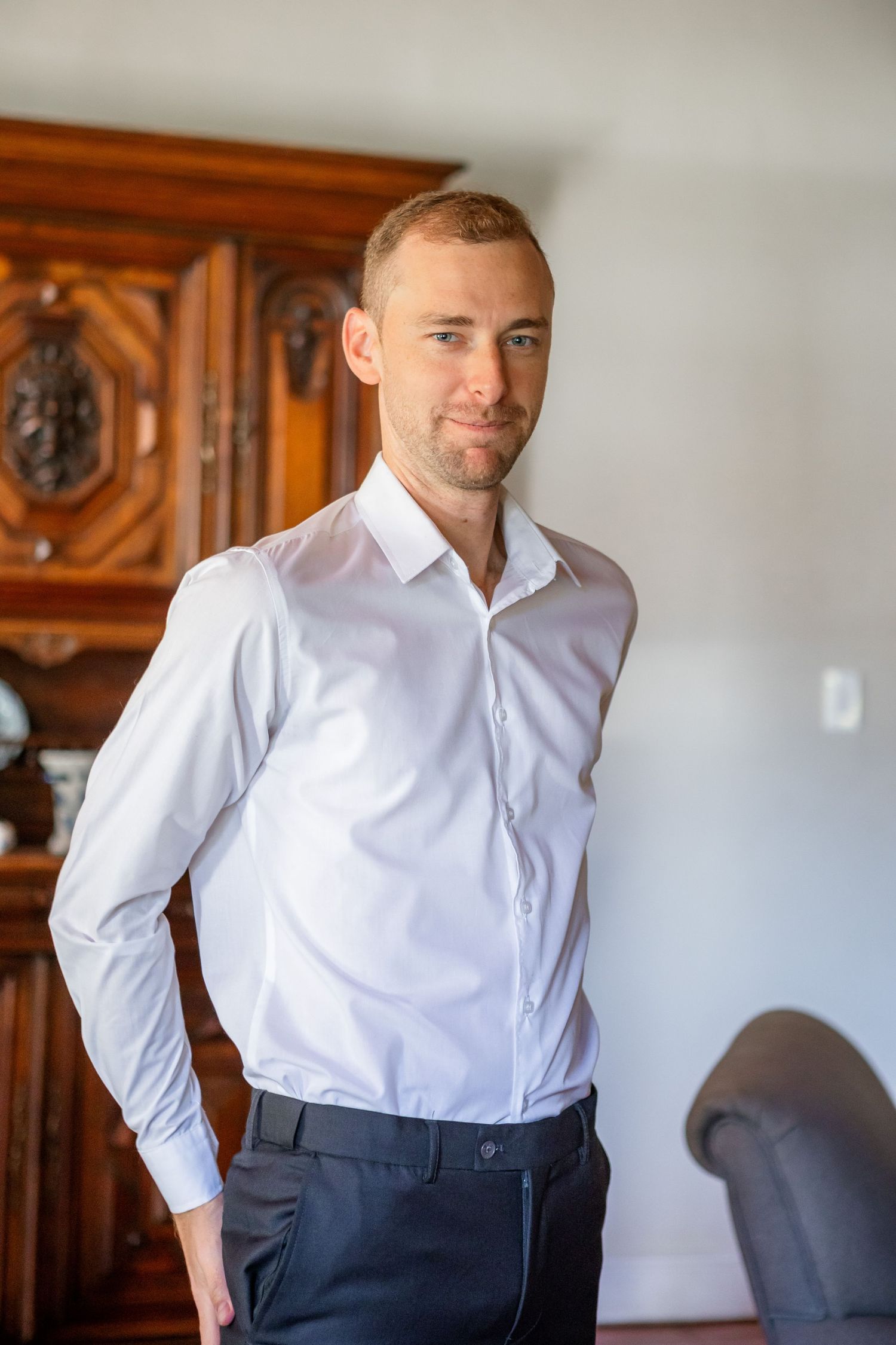 A person wearing a crisp white dress shirt and navy pants poses in a room with ornate wooden furniture.